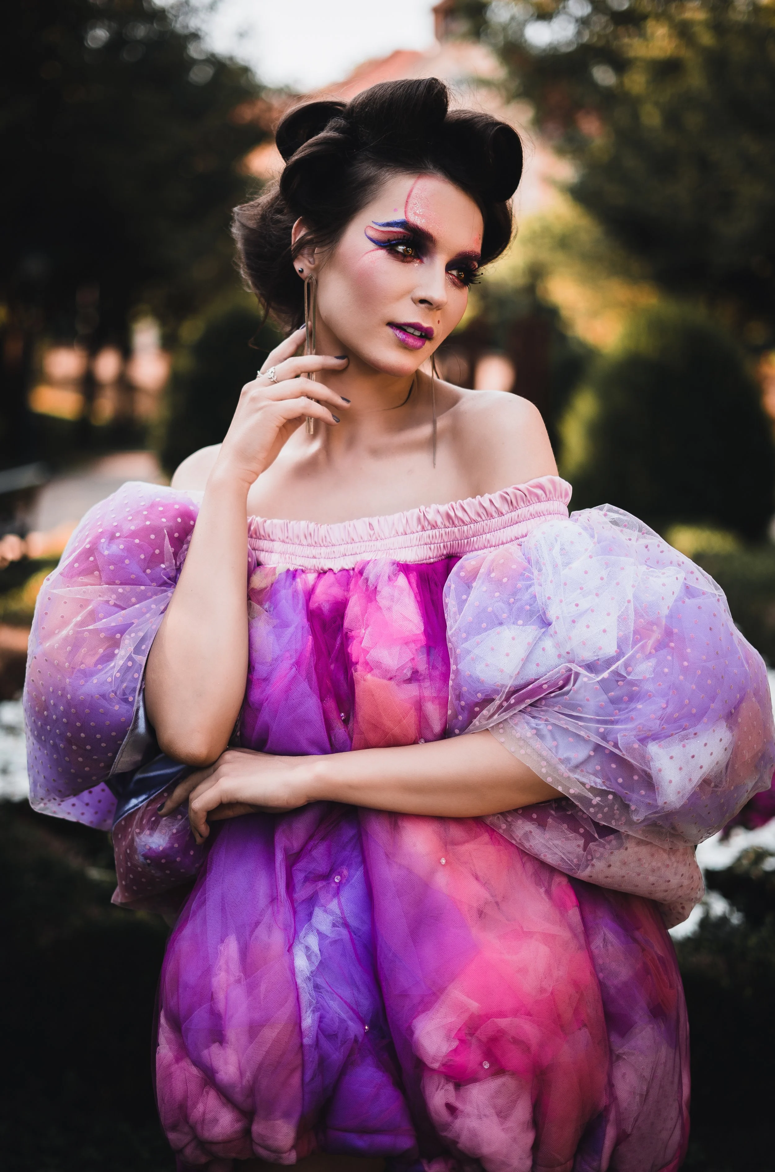 Junge Frau mit buntem Makeup und farbigem Kleid, im Freien bei Sonnenuntergang, mit Bäumen im Hintergrund.