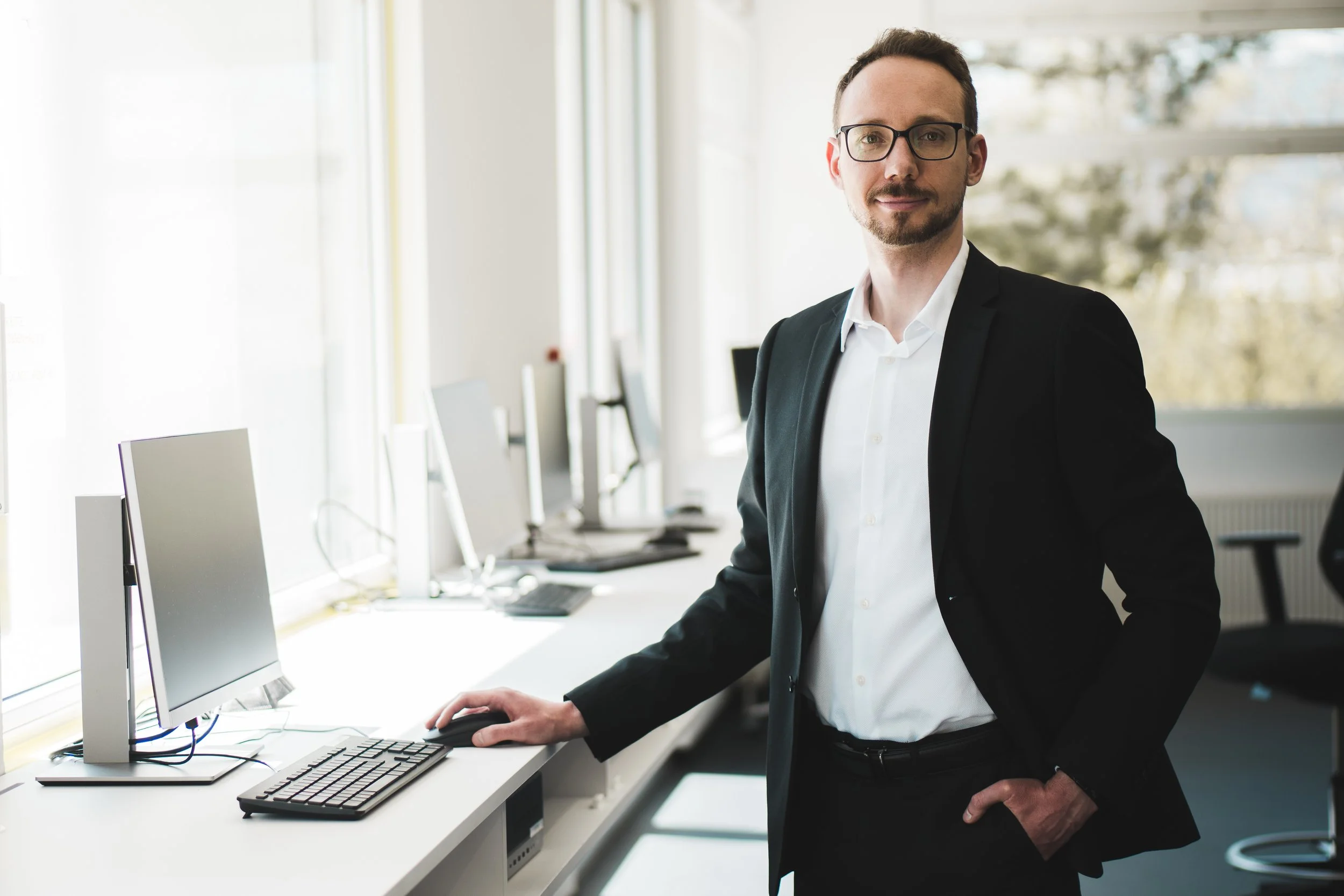 Ein Mann in formeller Kleidung steht an einem Computerarbeitsplatz in einem Büro, mit Computermonitor, Tastatur und Maus auf dem Schreibtisch.