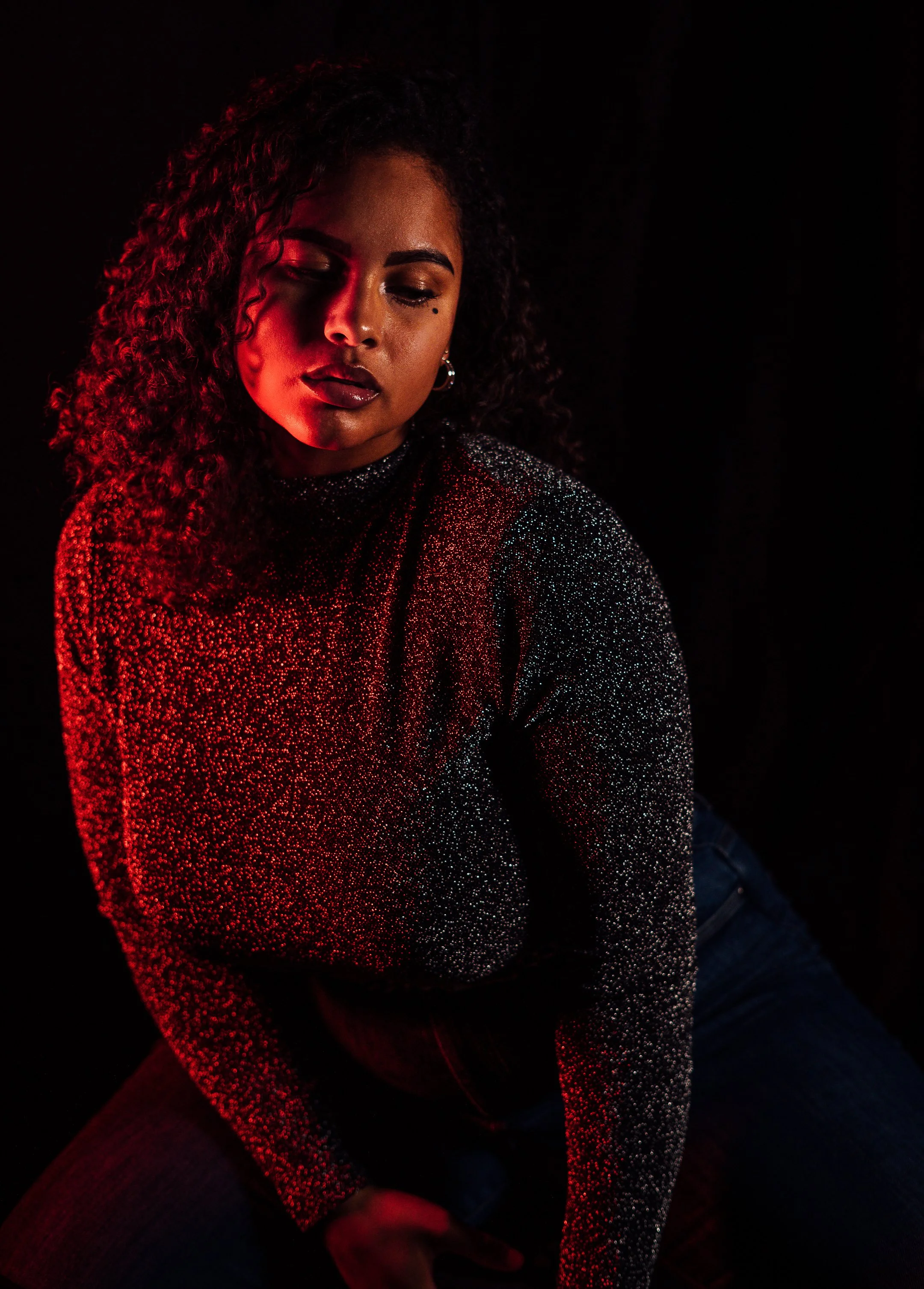 Junge Frau mit geschlossenen Augen, dunklem Lippenstift, und einem schwarzen Glitzerpullover, im Dunkeln. Rotes Licht beleuchtet sie.