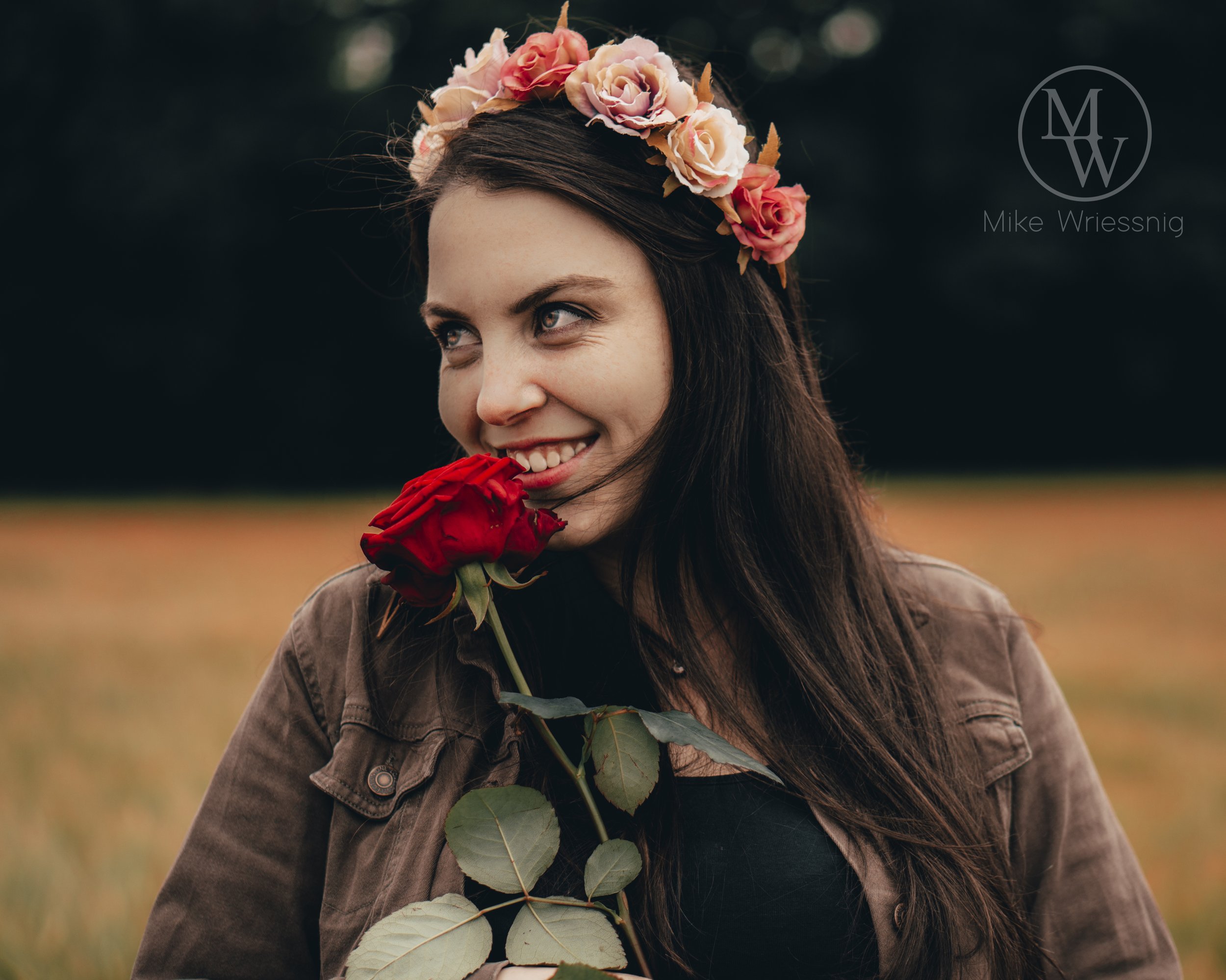 Junge Frau trägt einen Blumenkranz aus Rosen auf dem Kopf und hält eine rote Rose. Sie lächelt bei einem Spaziergang im Freien.