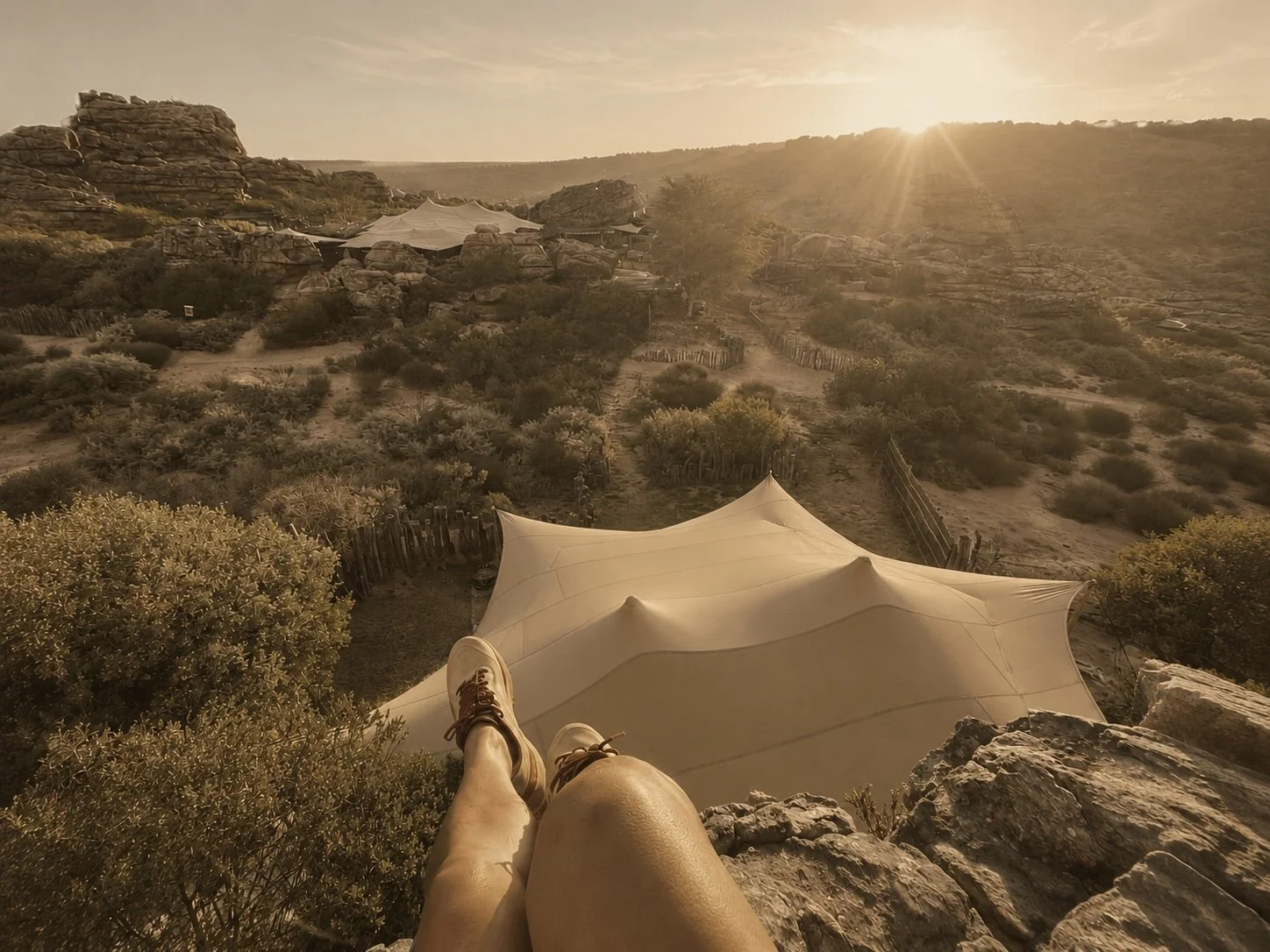 Person sitting on a rocky hill at sunset, overlooking a desert landscape with shrubs, rocks, and a white canopy tent in the distance.