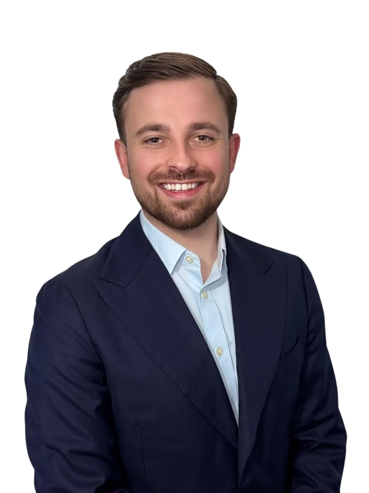 A young man with short brown hair, a beard, and a mustache, dressed in a dark navy blazer and a light blue collared shirt, smiling against a plain white background.