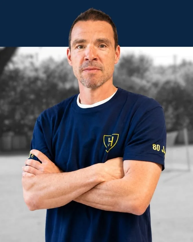 Mann mit kurzen dunklen Haaren und leichtem Bart, trägt ein dunkelblaues T-Shirt mit einem gelben Emblem, steht mit verschränkten Armen vor einem unscharfen Hintergrund.
