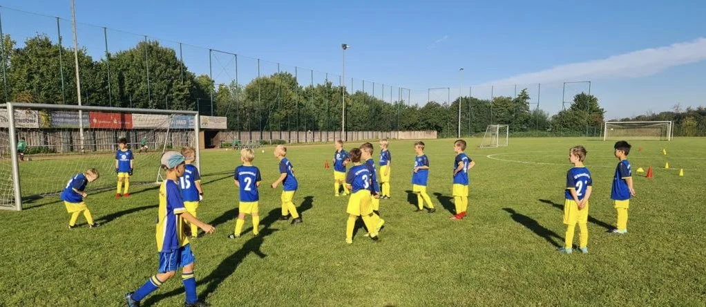 Junge Kinder in blauen Trikots und gelben Hosen spielen Fußball auf einem grünen Spielfeld an einem sonnigen Tag.