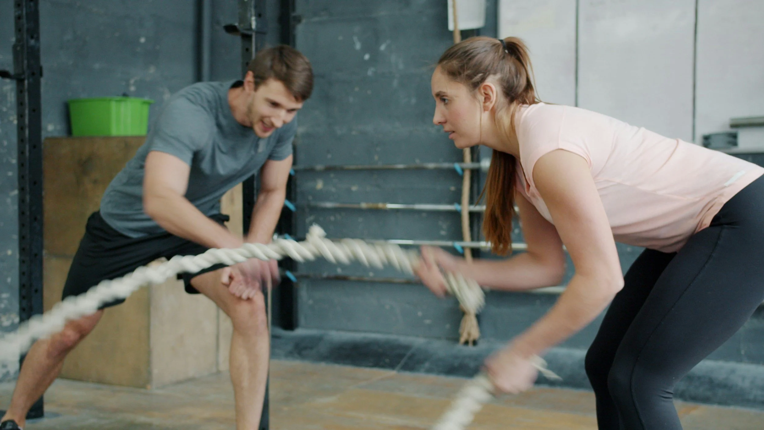 Zwei Menschen trainieren im Fitnessstudio, indem sie an einem Seil ziehen, in einer Haltung, die auf Krafttraining hindeutet.
