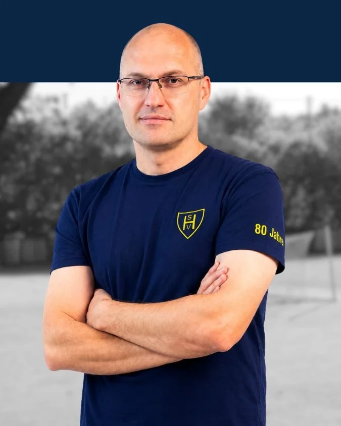 Mann mit Brille, kurzhaarig, in blauer T-Shirt mit Logo, steht mit verschränkten Armen vor doppeltem Hintergrund, farbig im Vordergrund, schwarz-weiß im Hintergrund.
