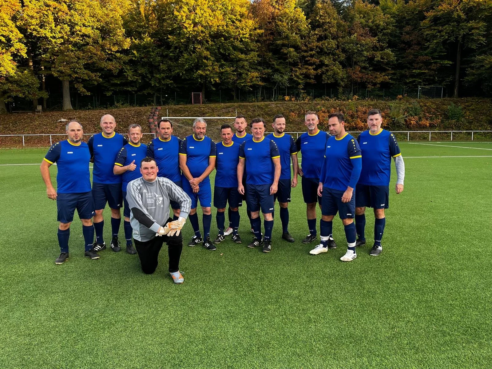 Ein Fußballteam in blauen Trikots auf einem Fußballfeld während des Tages, mit einemTrainer in grauem Trikot im Vordergrund, umgeben von Bäumen im Herbst.