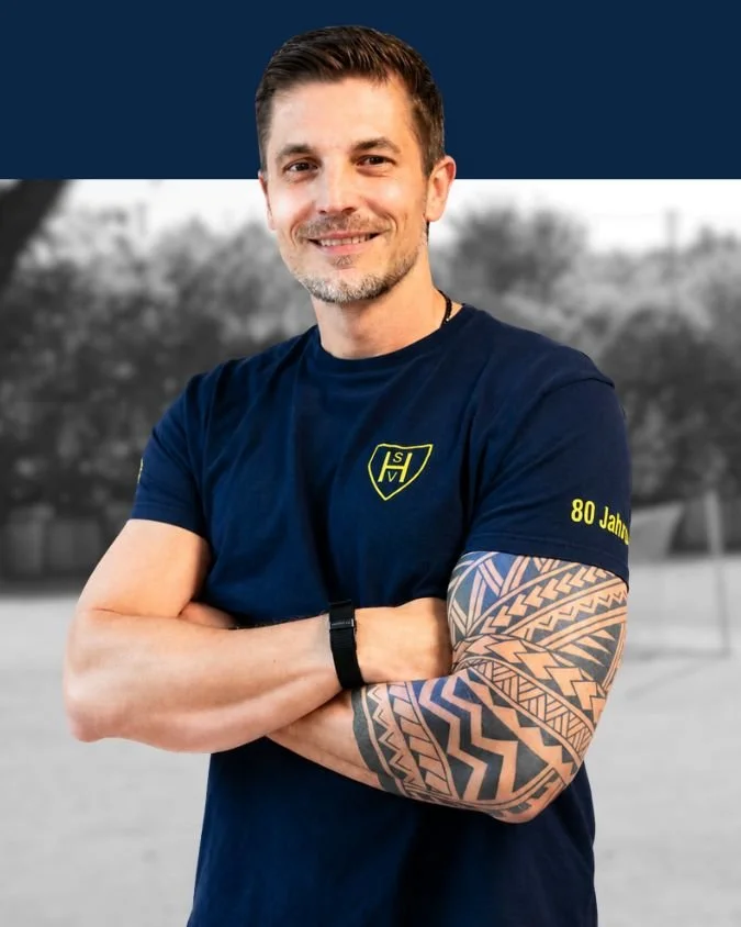 Mann mit kurzen dunklen Haaren, grauem Bart, trägt ein dunkelblaues T-Shirt mit einem Logo und schließt die Arme vor der Brust, zeigt sein Tattoo auf dem linken Arm.