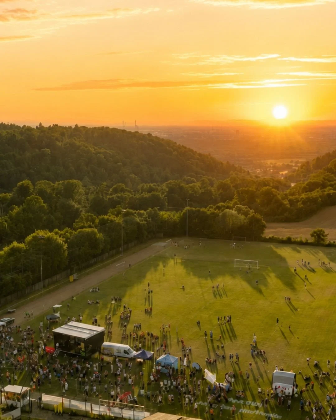 Sonnenuntergang über einem Sportplatz mit Menschen, Ständen und Tieren, umgeben von Grünflächen und Hügeln.