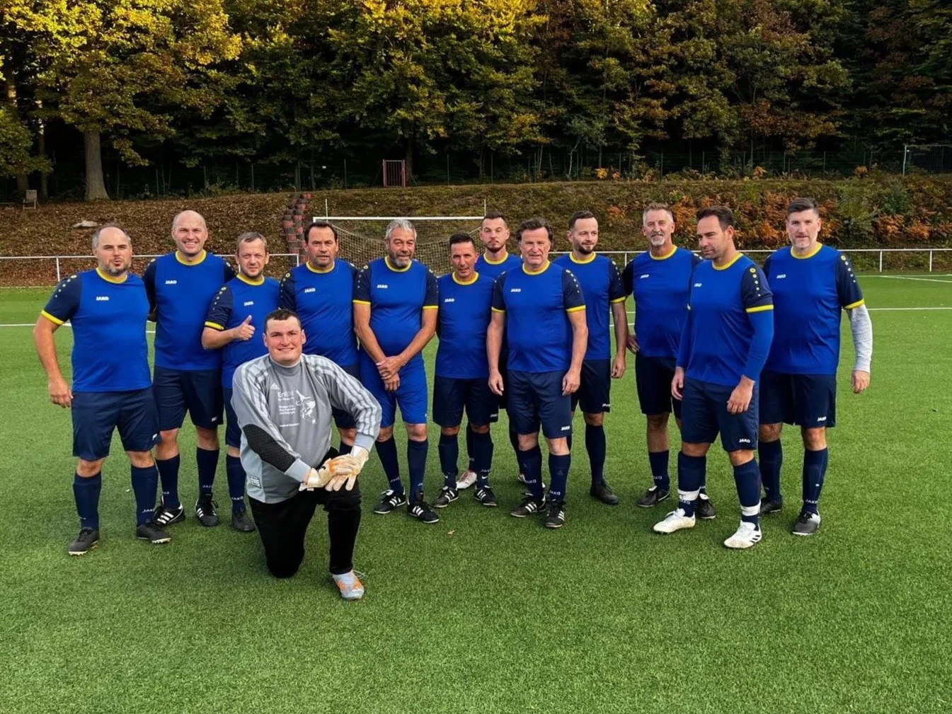 Eine Gruppe von zwölf Männer in blauen Fußballtrikots mit gelben Akzenten steht auf einem Fußballfeld. Einer der Männer trägt einen Torwart-keeper-Overall. Im Vordergrund ist ein Mann in grauem Torwart-Overall, der auf einem Knie sitzt, und lächelt. Im Hintergrund sind Bäume mit herbstlicher Laubfärbung zu sehen.