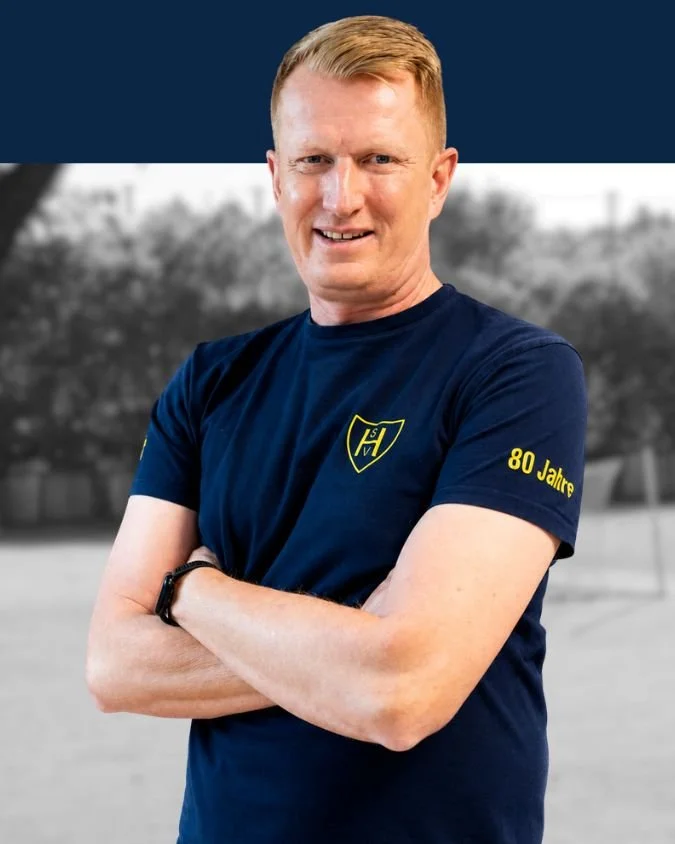 Mann in blauer T-Shirt mit gelbem Logo und der Aufschrift "80 Jahre" auf dem rechten Ärmel, Arme verschränkt, im Hintergrund Bäume, unscharf, Outdoor