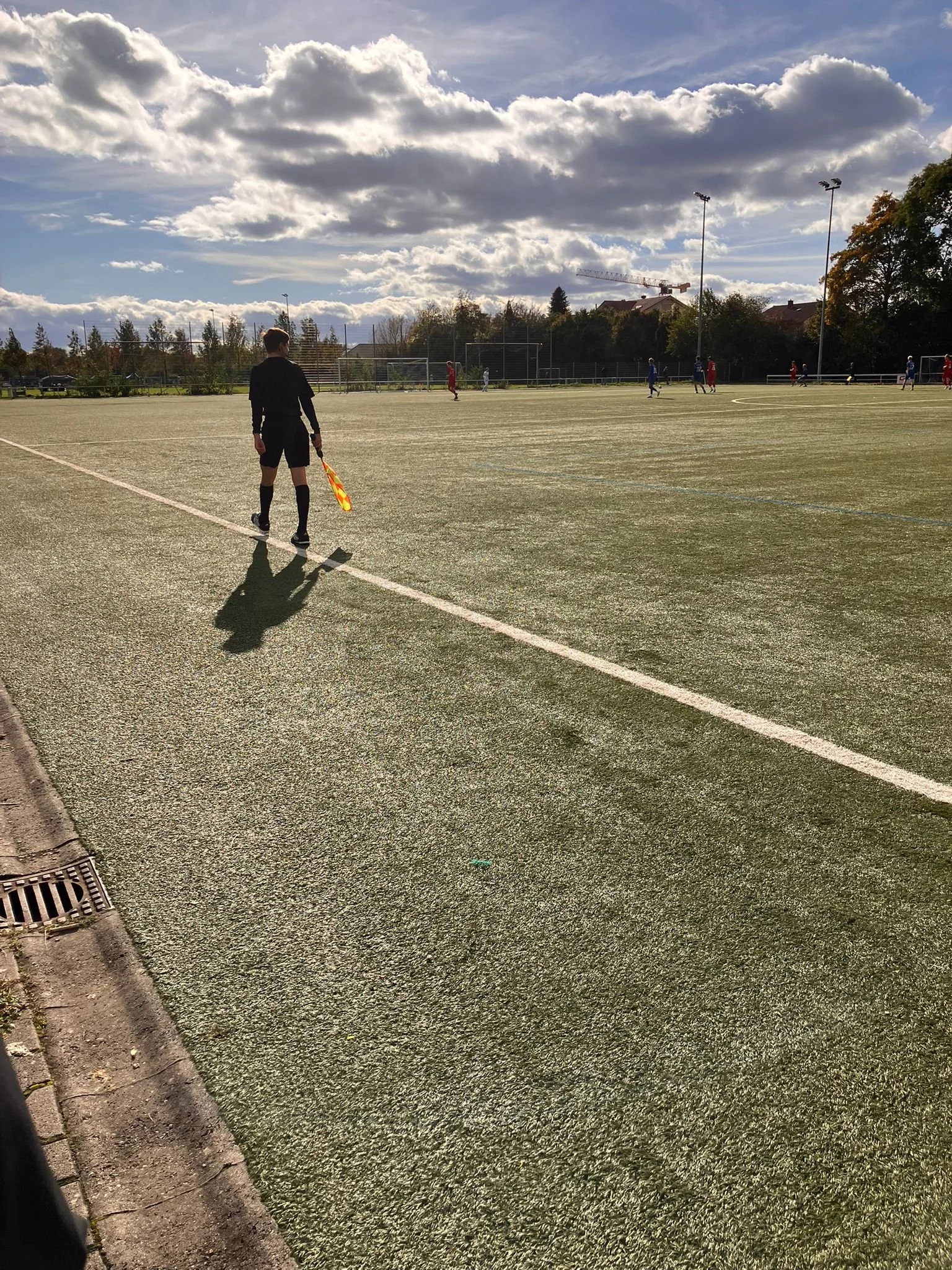 Ein Fußballspielfeld bei Tageslicht mit einem Schiedsrichter am Spielfeldrand und mehreren Spielern im Spiel, im Hintergrund Bäume und eine Baukran.