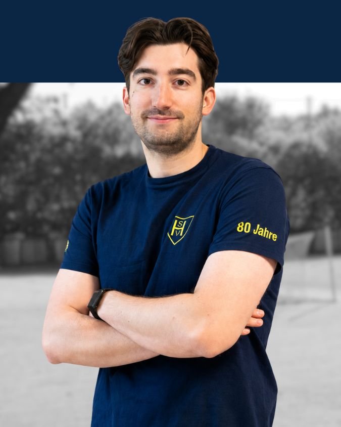 Junger Mann mit braunen Haaren trägt ein blaues T-Shirt mit einem Logo und der Aufschrift "80 Jahre" am rechten Ärmel, steht vor einer unscharfen Naturkulisse, Arme verschränkt, lächelnd.