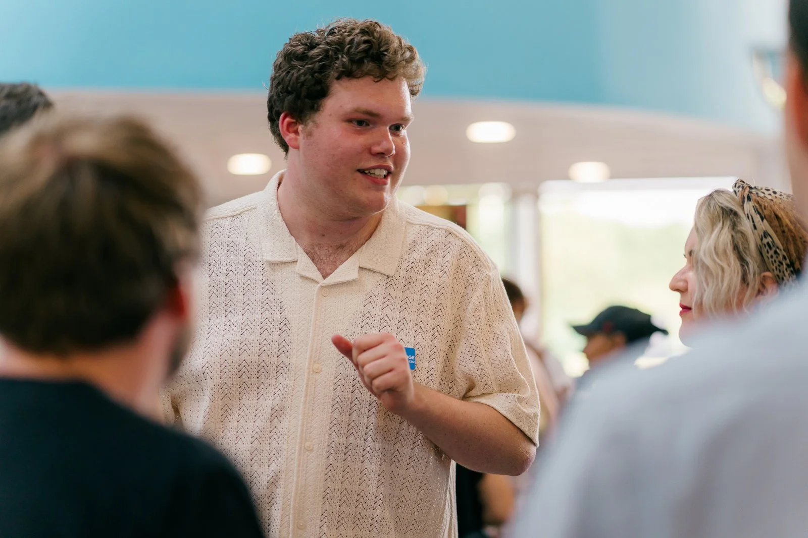 A young man with curly hair speaking to a group of people at an indoor event, wearing a beige short-sleeved shirt, with a woman with blonde hair and a patterned headband listening to him.