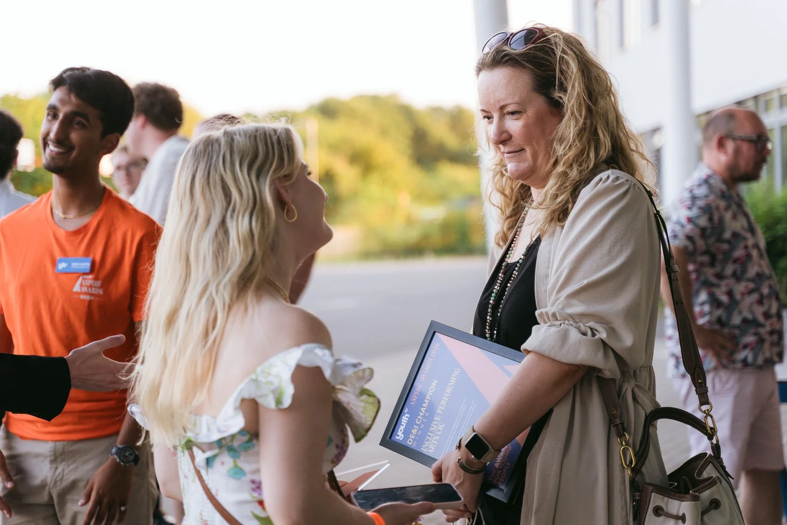A woman handing a certificate to a young girl at an outdoor event, with other people in the background.
