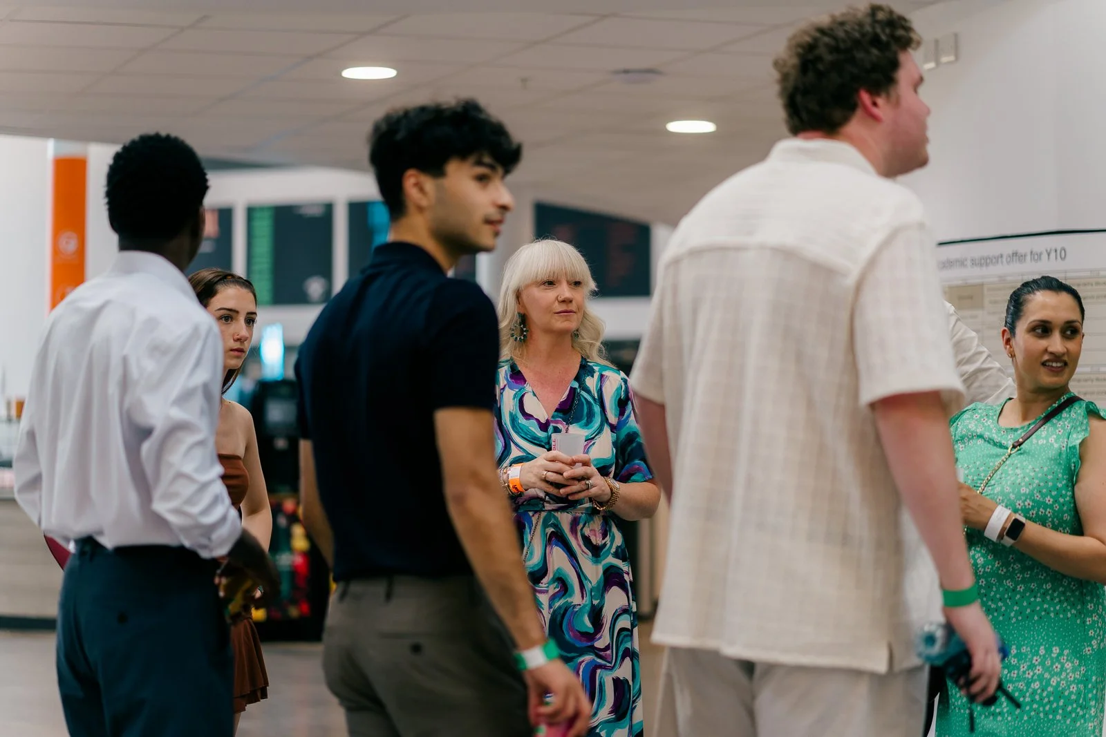 Group of six diverse people standing and talking at an indoor event or conference, some holding drinks, with screens and posters in the background.