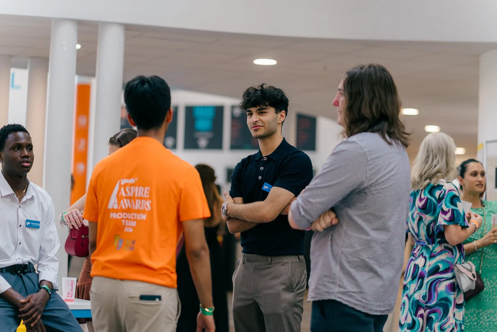 A group of people gathering in a modern indoor space, including a man in black with arms crossed talking to a woman with brown hair wearing a gray striped shirt, and a person in an orange shirt with 'Aspire Awards Production Team 2022' printed on the back, during an event or conference.