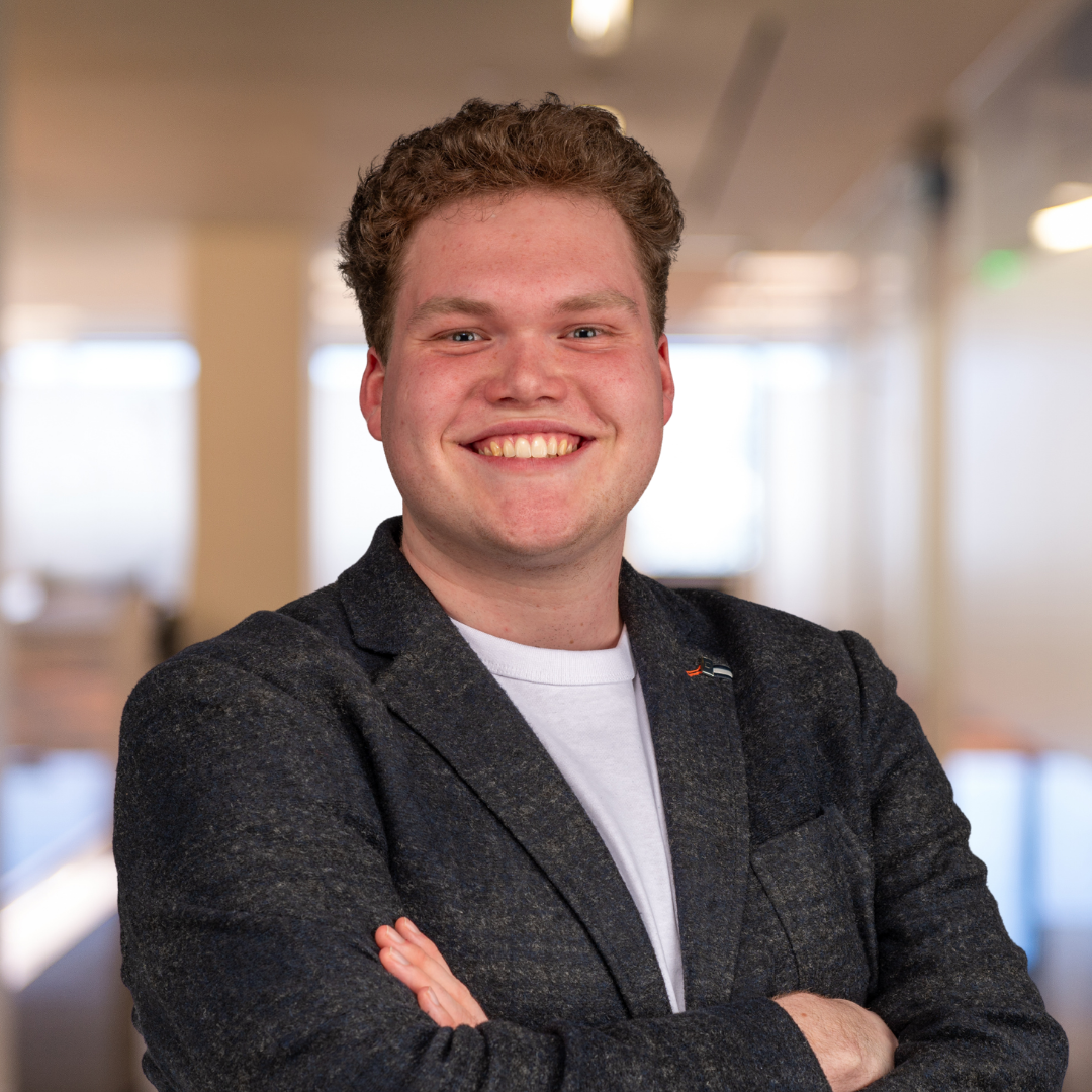 Portrait of a young man with curly hair, smiling, dressed in a blue blazer and light blue shirt, against a blue gradient background.