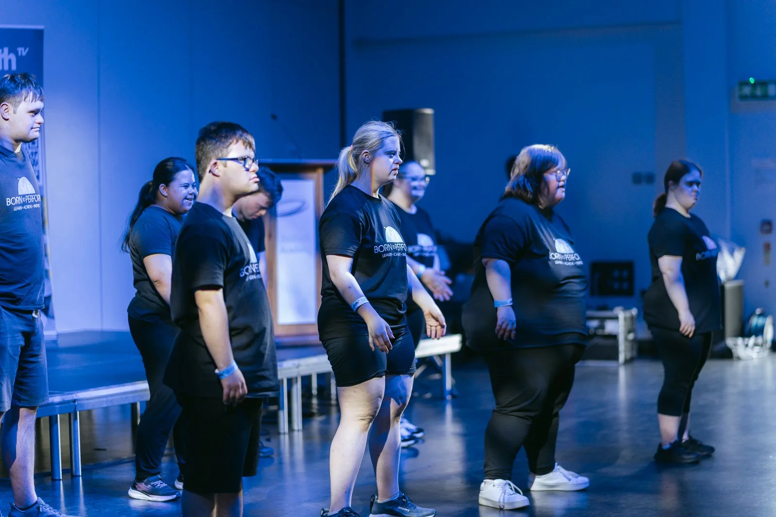 Group of diverse people standing in a theater or auditorium, participating in an activity or rehearsal, wearing matching black t-shirts with a logo, illuminated by blue stage lighting.