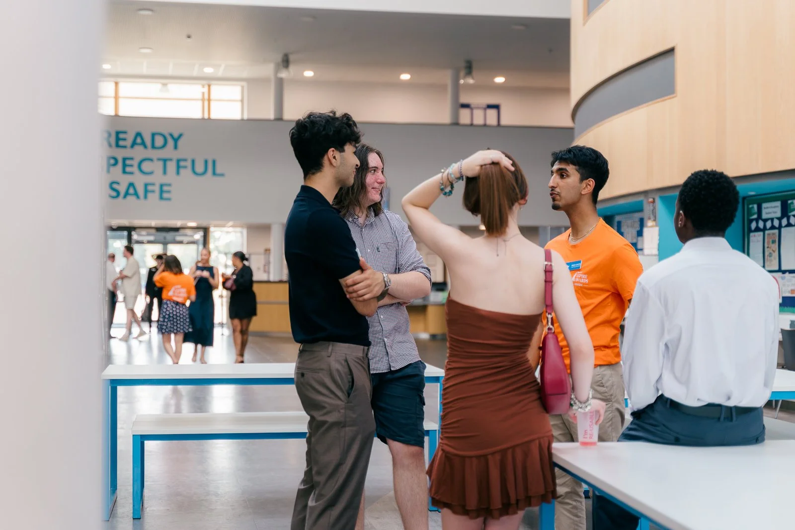 Group of five diverse young adults conversing in a modern, well-lit public space, with some people in the background walking or talking.