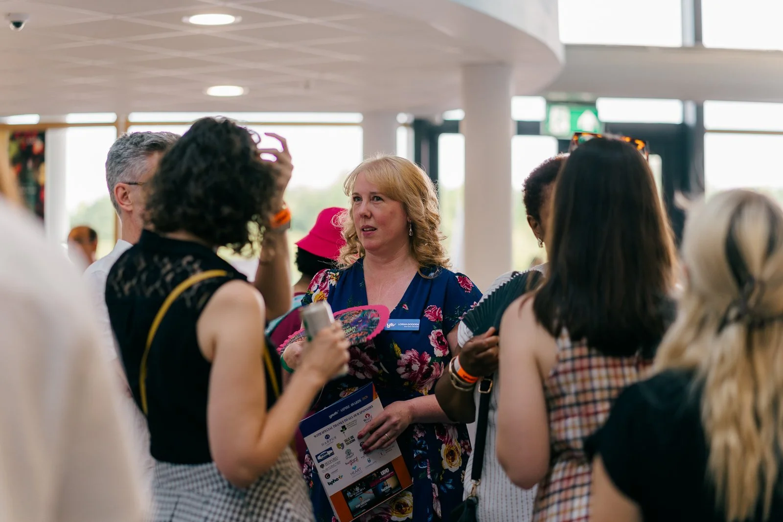 Group of diverse people talking and interacting indoors, one woman in a floral dress holding a brochure, others listening and engaging in discussion.