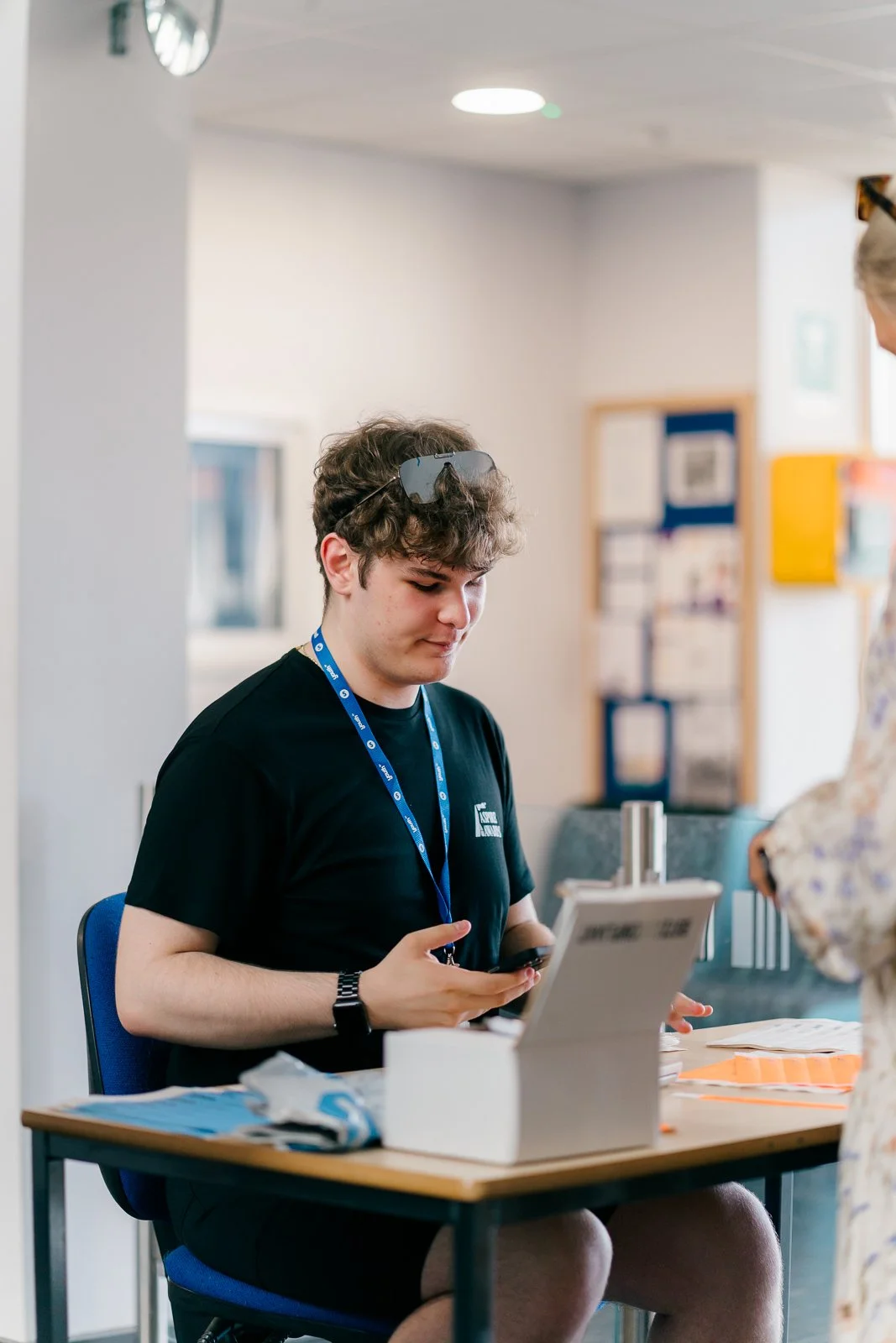 A young man with curly hair, wearing a black t-shirt, sitting at a registration desk with a lanyard around his neck, looking at his phone, with sunglasses resting on his head. There is a woman standing in front of him at the desk.