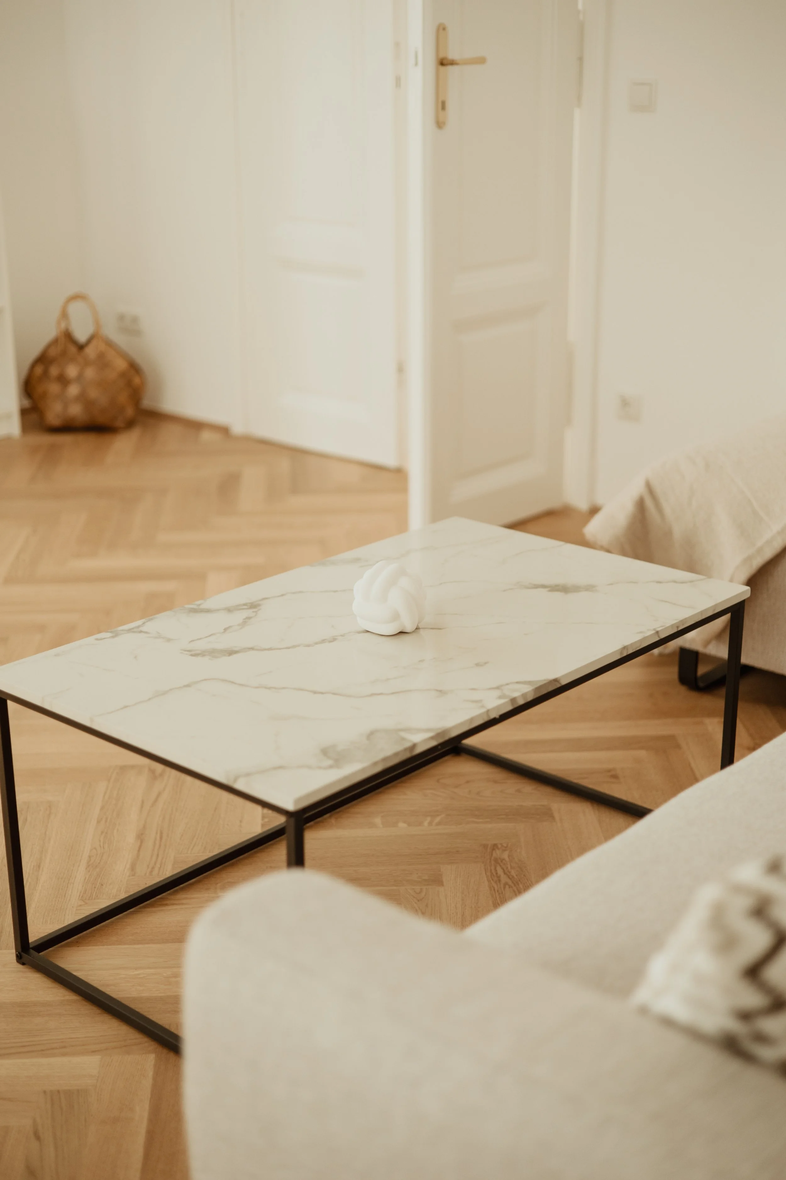 Ein Wohnzimmer mit einem weißen Sofa, einem Couchtisch mit Marmorplatte und einer weißen Wand. Im Hintergrund eine Tür und eine Tasche auf dem Boden.