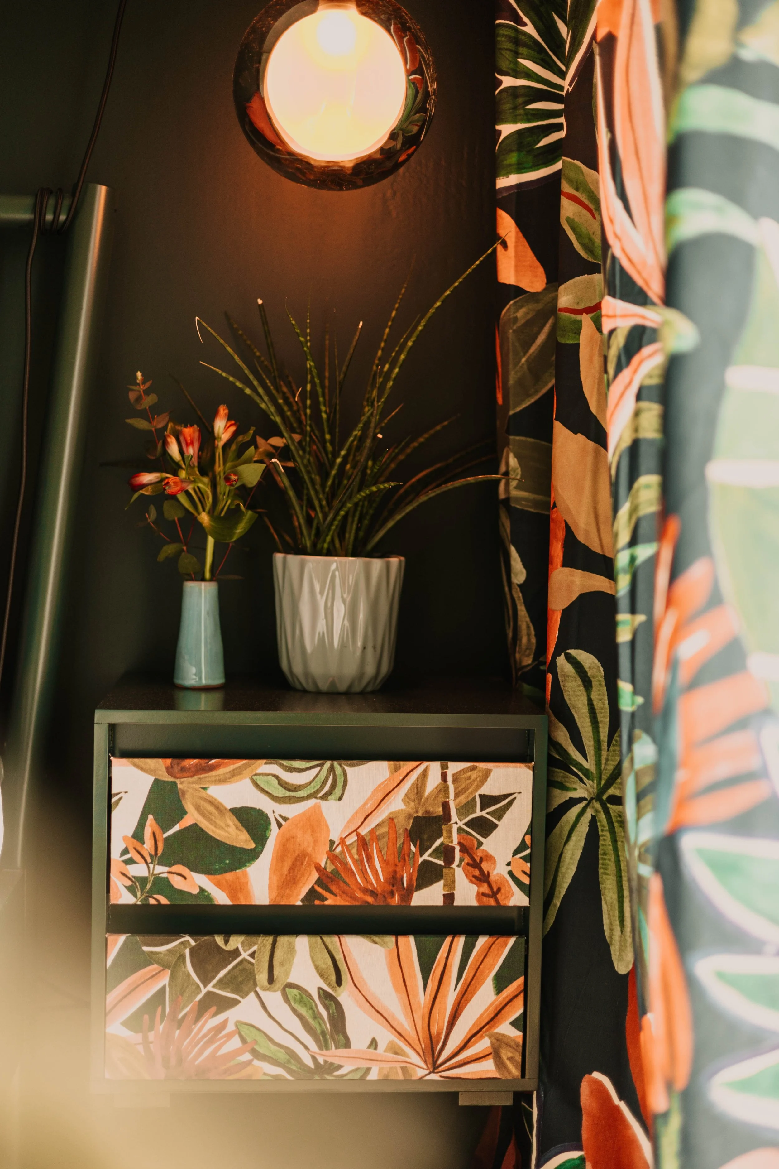 Ein beleuchteter Raum mit einer Vase mit grünen und roten Pflanzen auf einem Regal, daneben eine Schublade mit sommerlichem Blumenmuster, und bunten Vorhängen mit Blattmotiven.