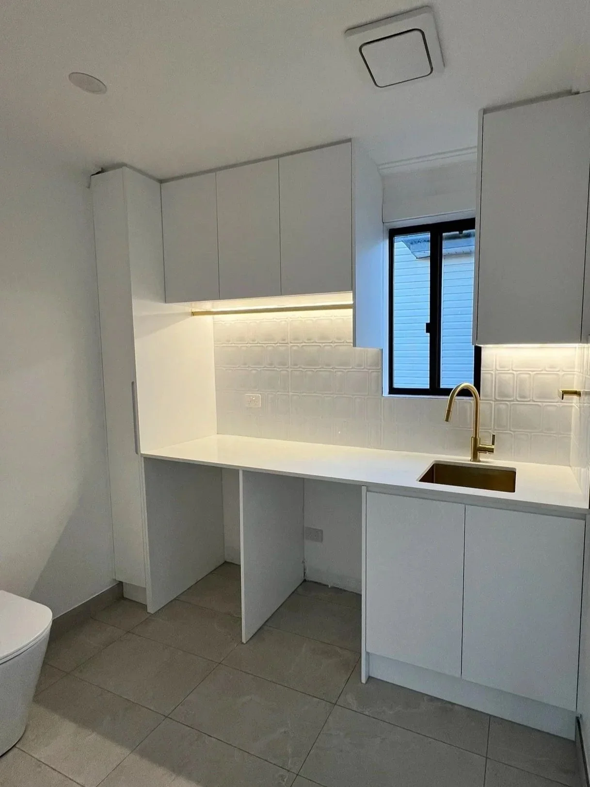 Modern white kitchen with gold faucet, white cabinets, and a small window with house siding visible outside.