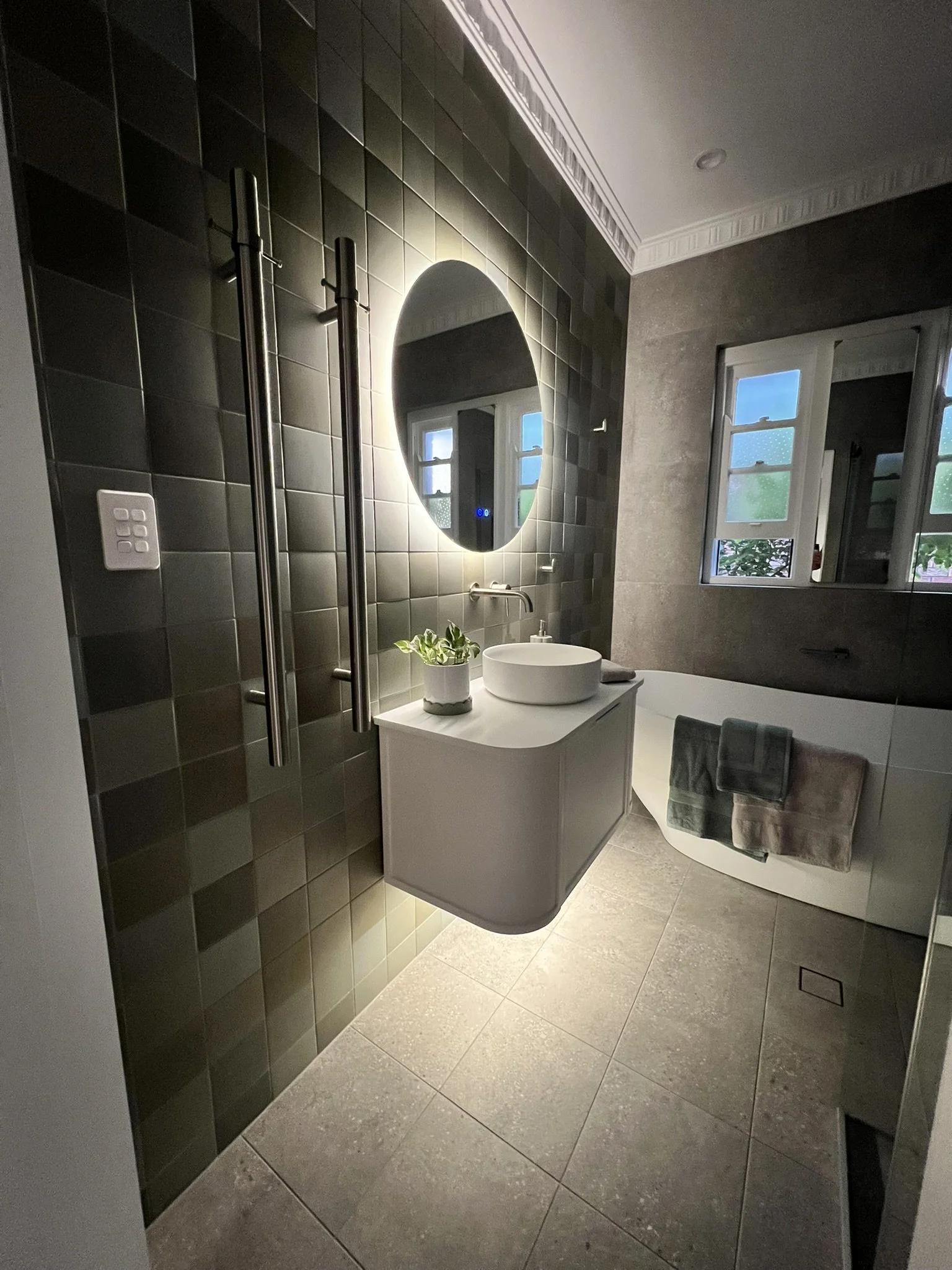 A modern bathroom with dark tiled walls, a large backlit oval mirror, a white sink with a potted plant on top, and a bathtub with a gray towel hanging over its edge. There is a window with two small windows showing a cloudy sky outside.