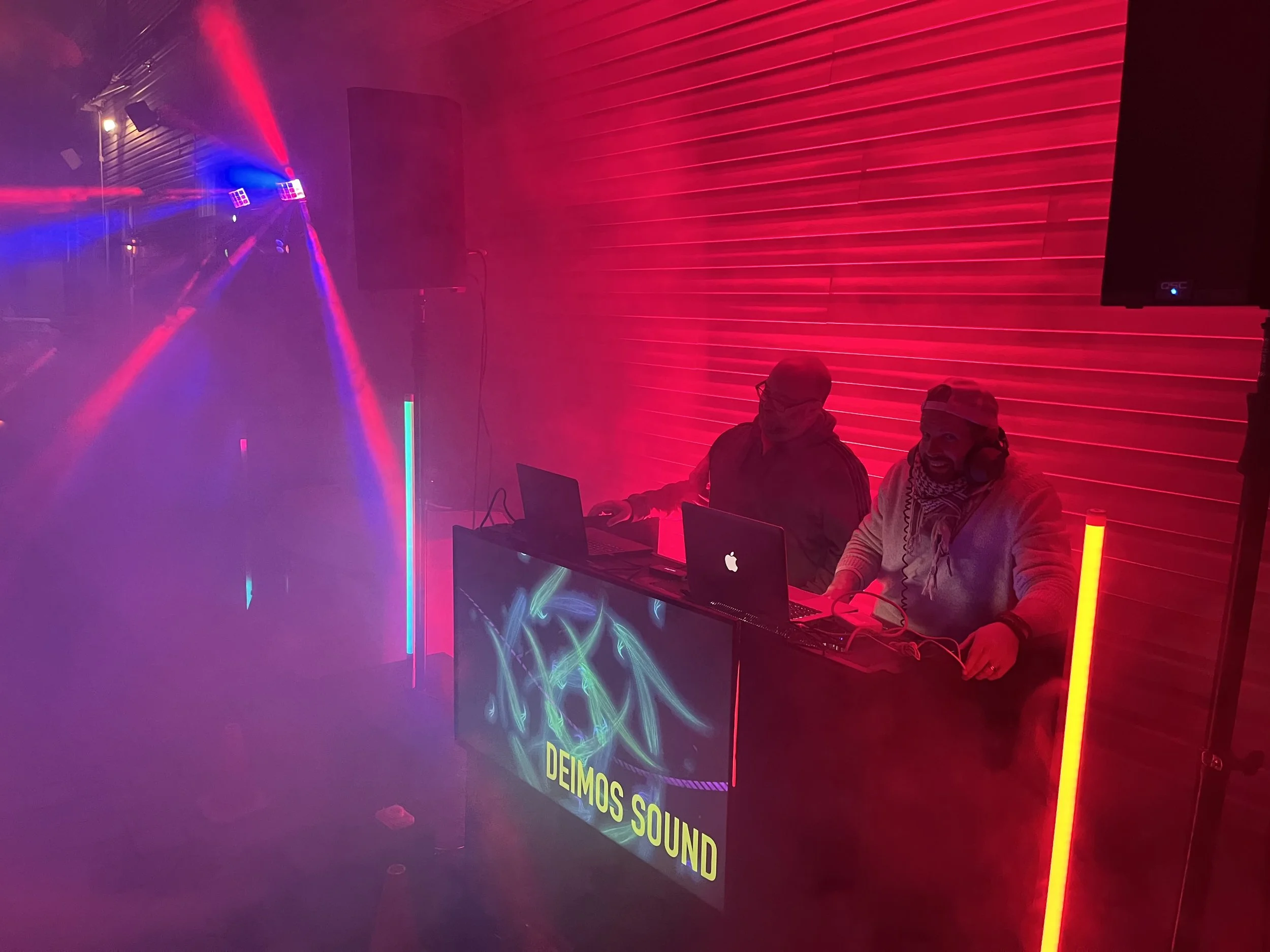 Two people sitting at a table, working on laptops, in a room with red lighting and laser light effects, with a neon sign indicating 'DEIMOS SOUND'.