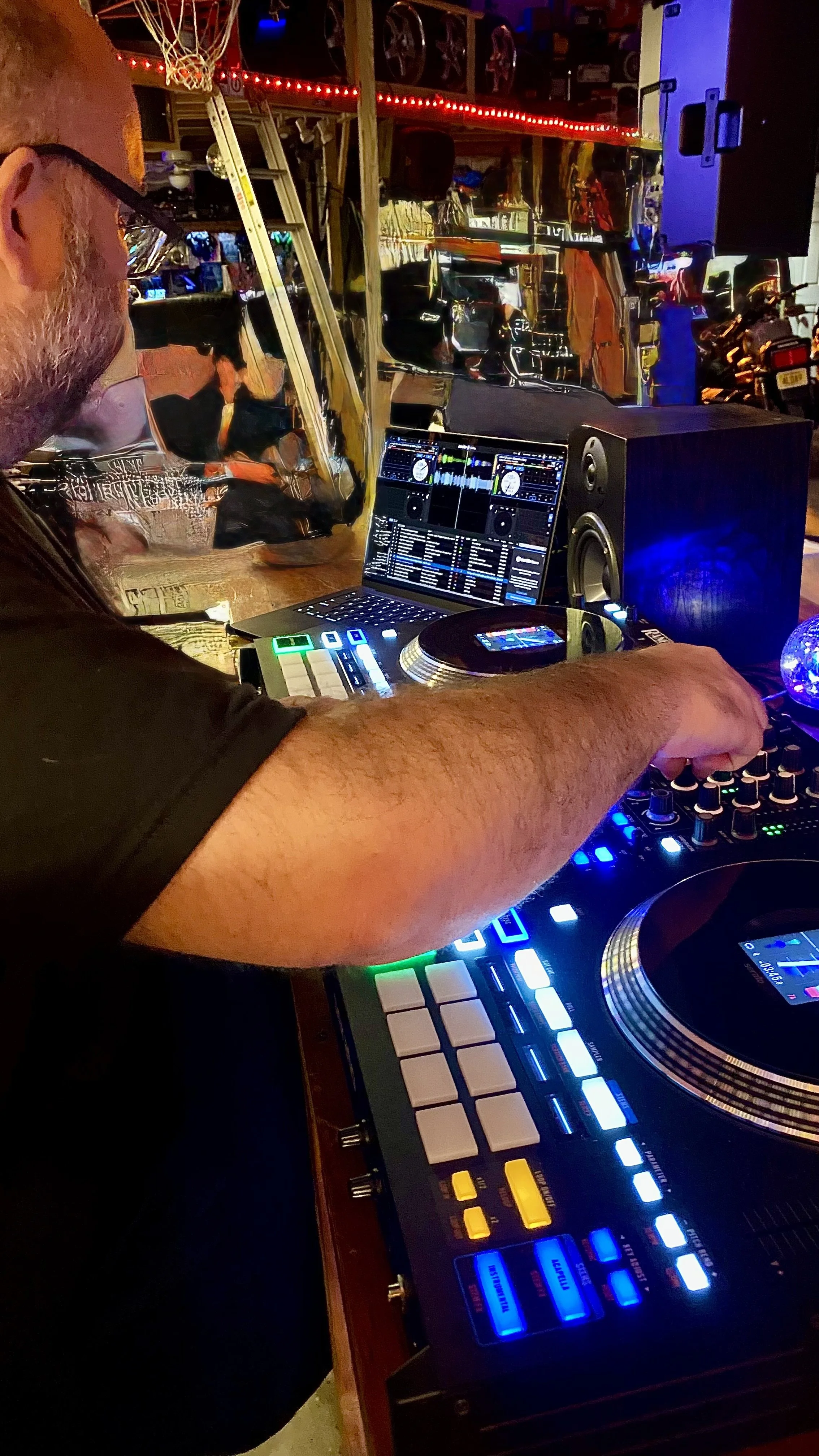 A DJ is performing with a DJ controller, laptop, and speaker in a dimly lit setting with colorful lights. The DJ is wearing glasses and has a beard, and is adjusting controls on the DJ equipment.