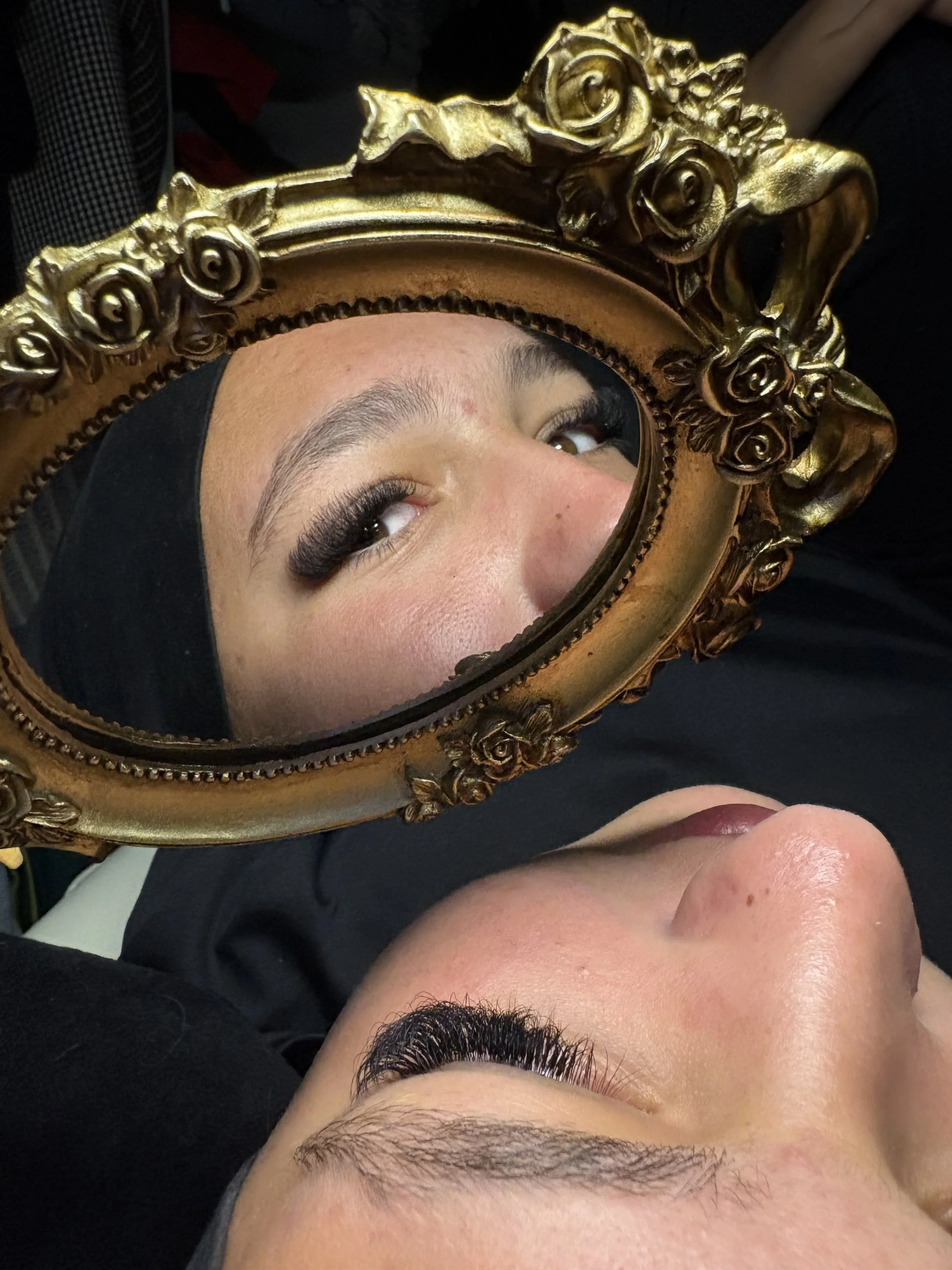 Deux personnes se regardent dans un miroir doré orné de roses, avec des maquillages de cils longs et épais.
