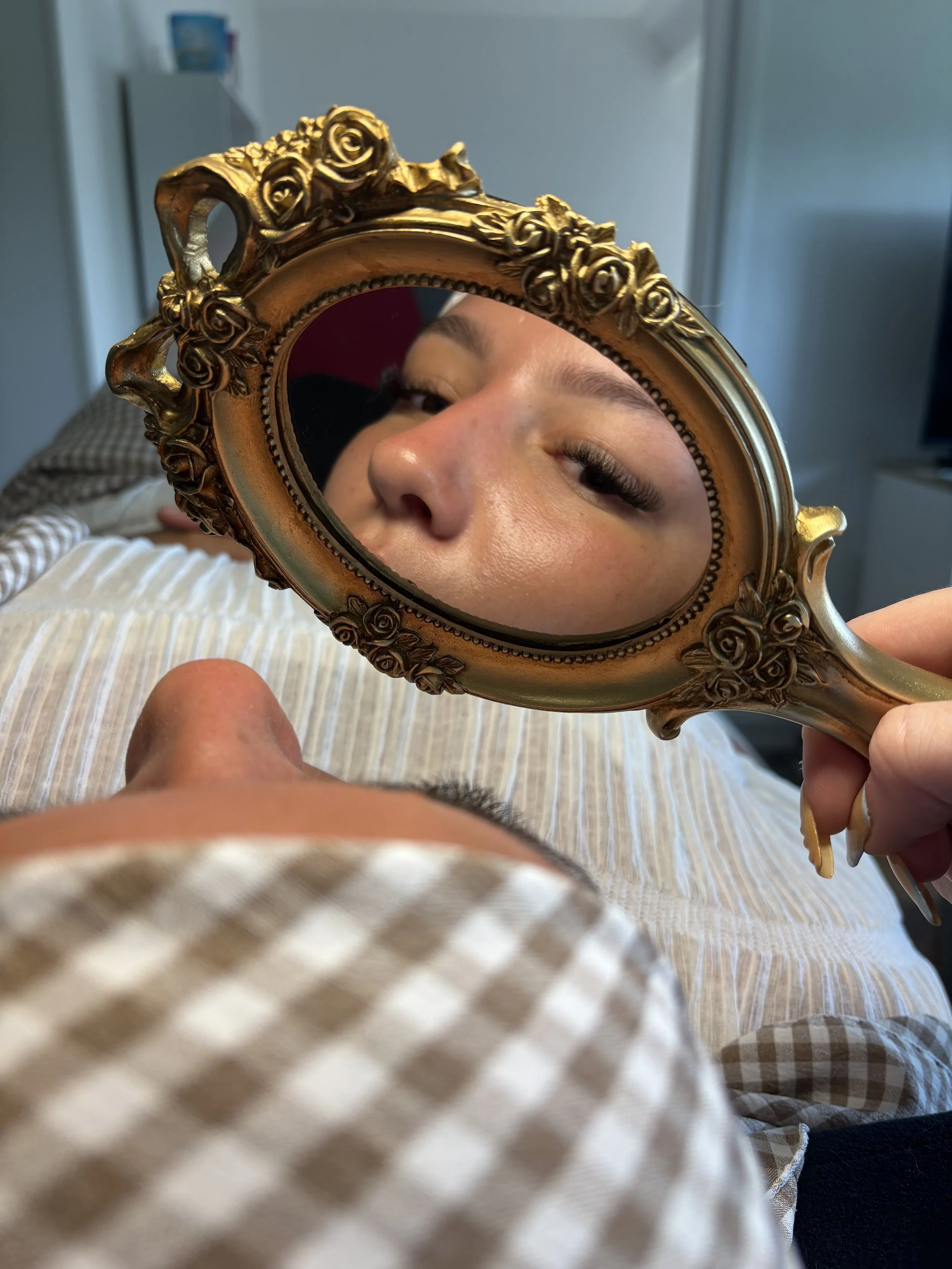 Une personne regarde son visage dans un miroir ovale avec un cadre doré orné de roses, tenu par une main. La photo est prise à partir d'une position sur le lit, avec une couverture rayée en premier plan et un mur gris en arrière-plan.