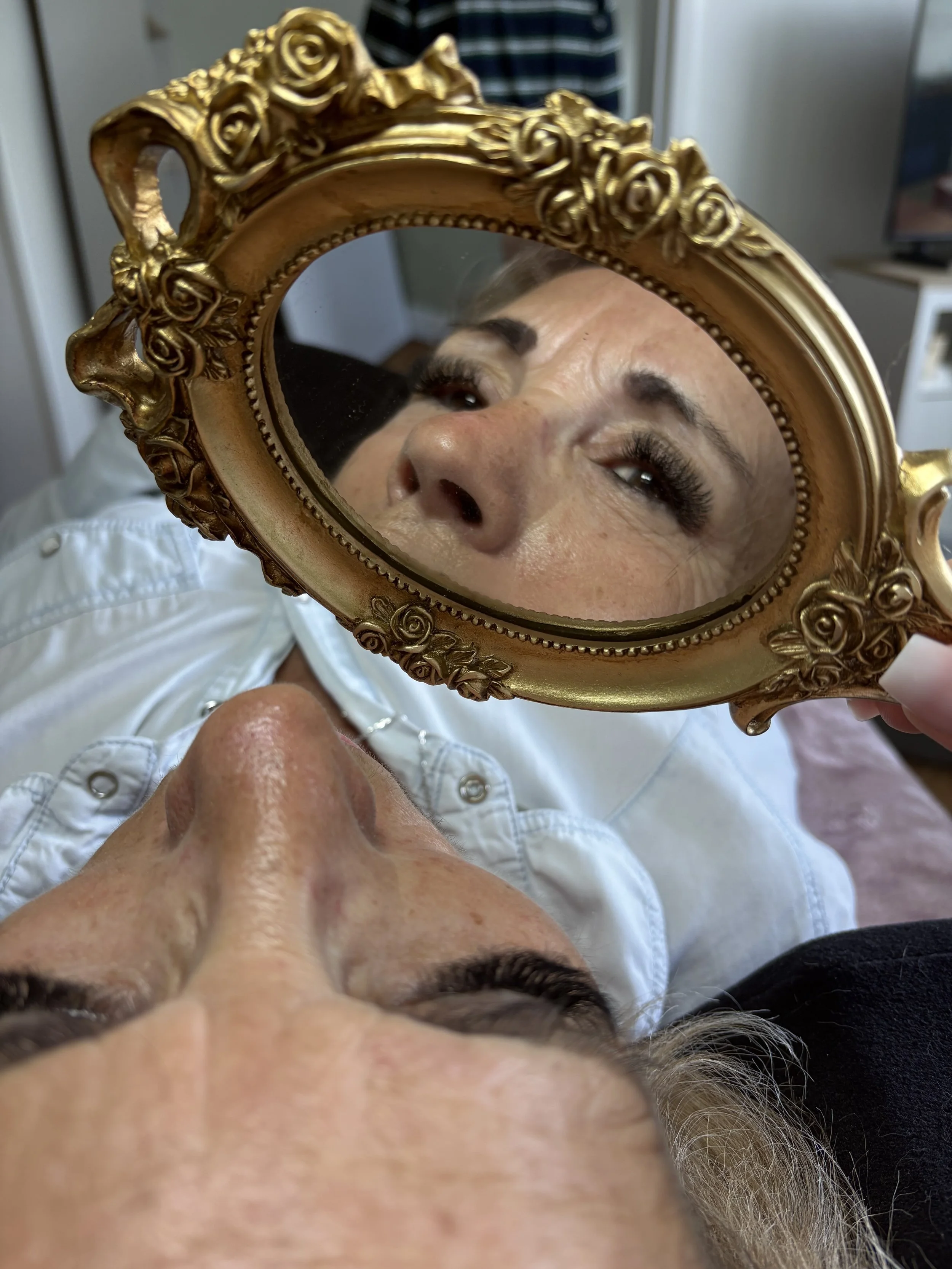 Un miroir doré à ornements floraux reflète le visage d'une femme regardant vers le dessus, avec des détails visibles de son visage et de ses yeux. La photo est prise en position inversée.