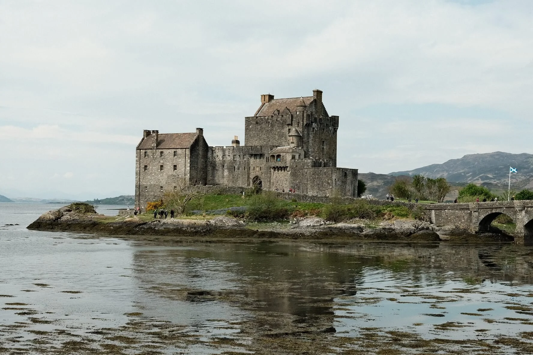 A historic castle with stone walls and towers, located on a small island surrounded by water, with mountains in the background and a Scottish flag flying nearby.