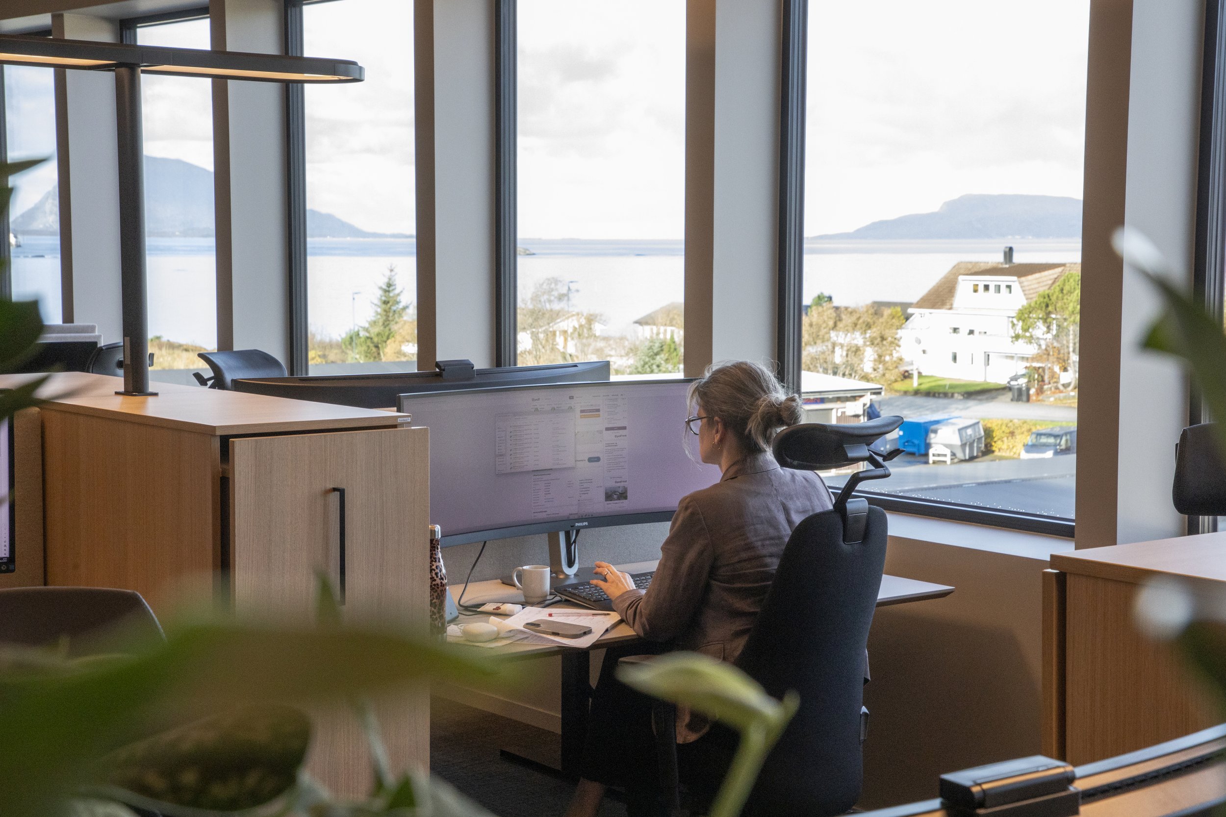 En kvinne som jobber på en kontorarbeidsplass med store vinduer som viser en utsikt over vann og fjell i bakgrunnen, med noen hytter og hus i nærheten.