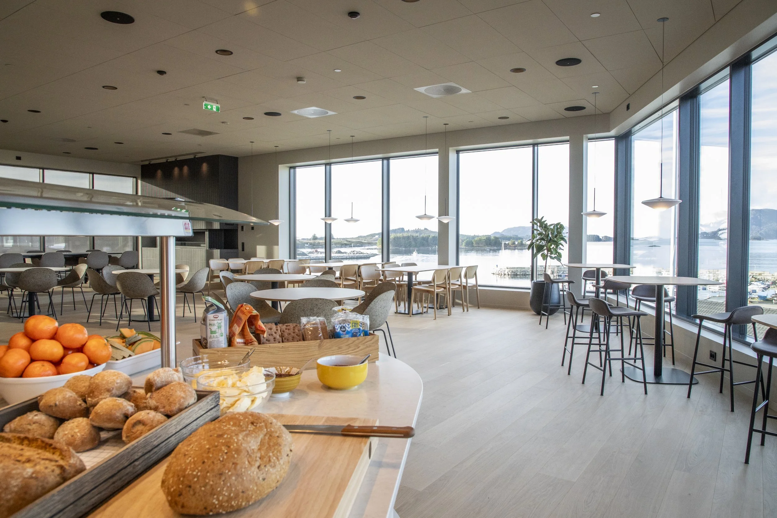 Et spiseområde med store vinduer som viser en vannutsikt, flere bord og stoler, og en frokostbuffé med brød og frukt i forkant.