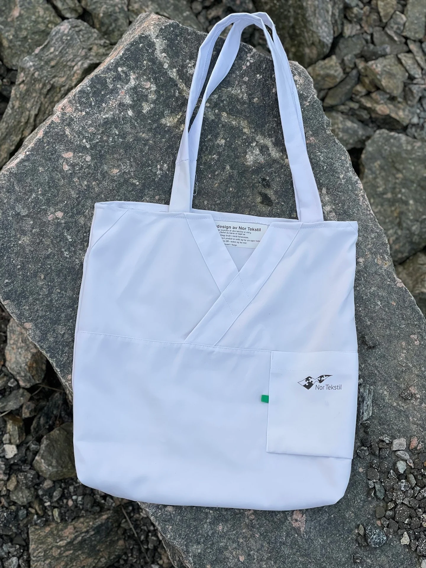 En hvit stoffpose med logo til Nor Tekstil ligger på en stor, flat stein med mindre steiner rundt.