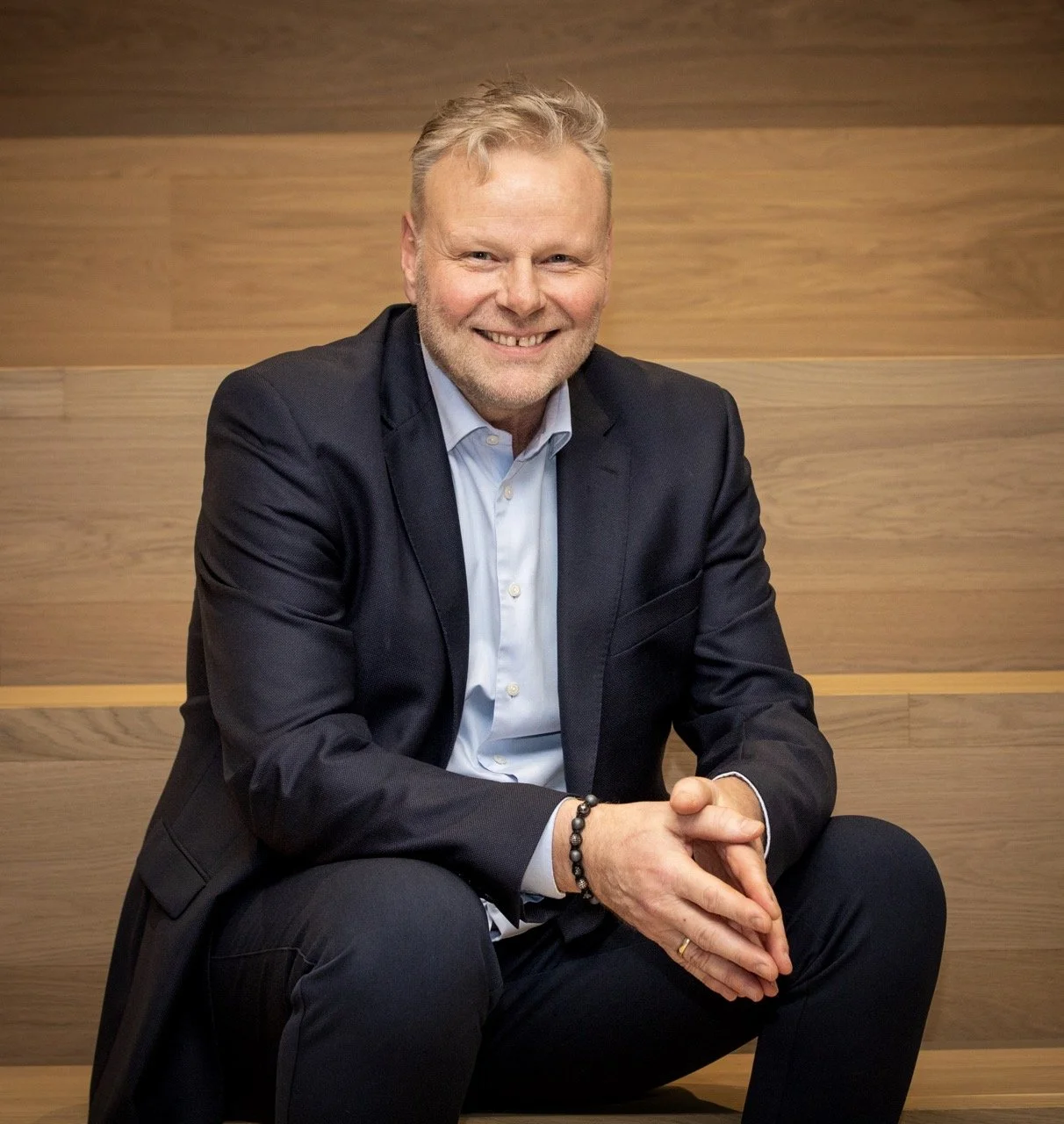 En mann i dress sitter mot en vegg av trepanel, smiler og har kort, lyst hår.