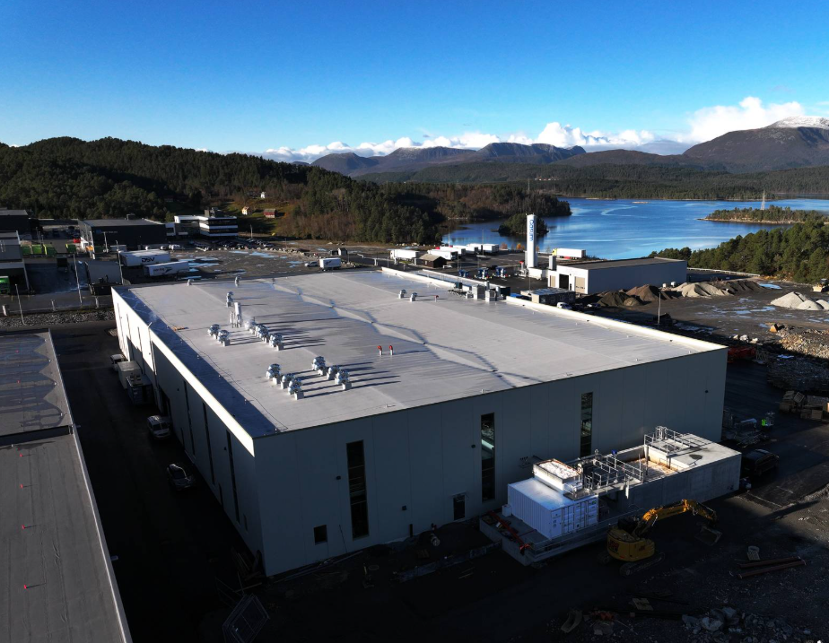 Et industriområde med byggearbeid, store hvite bygninger, en overskyet innsjø og fjell i bakgrunnen.