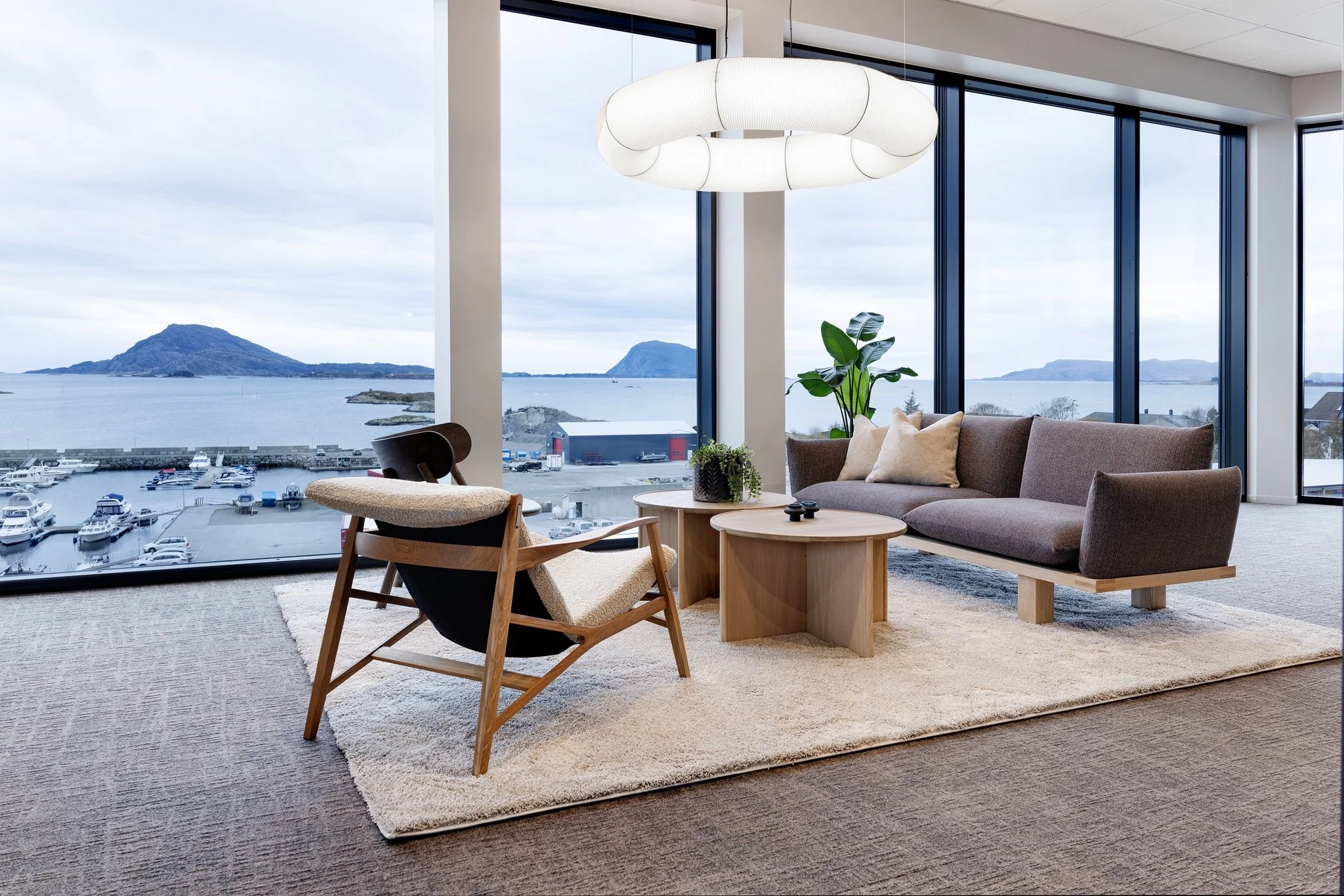 Innendørs stue med store vinduer med utsikt over hav og øyer, moderne lyse møbler, plante, og taklampe.