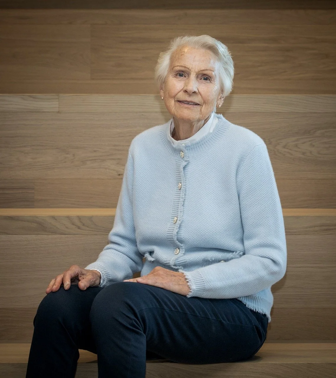 En eldre kvinne med hvitt hår, iført en lys blå genser og mørke bukser, sitter på en trebord med en trevegg i bakgrunnen.