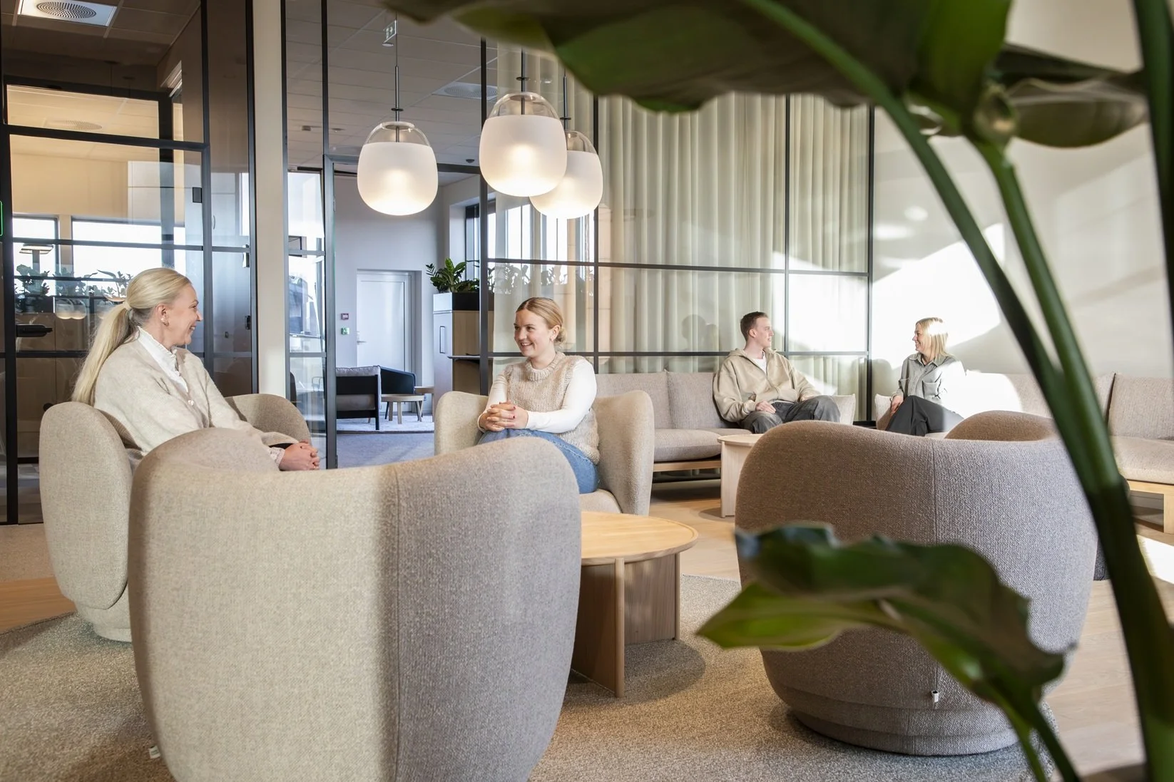 Fire mennesker i en moderne lounge, to kvinner i forgrunnen som snakker og ler, to personer i bakgrunnen som prater, omgitt av komfortable stoler og myk belysning.