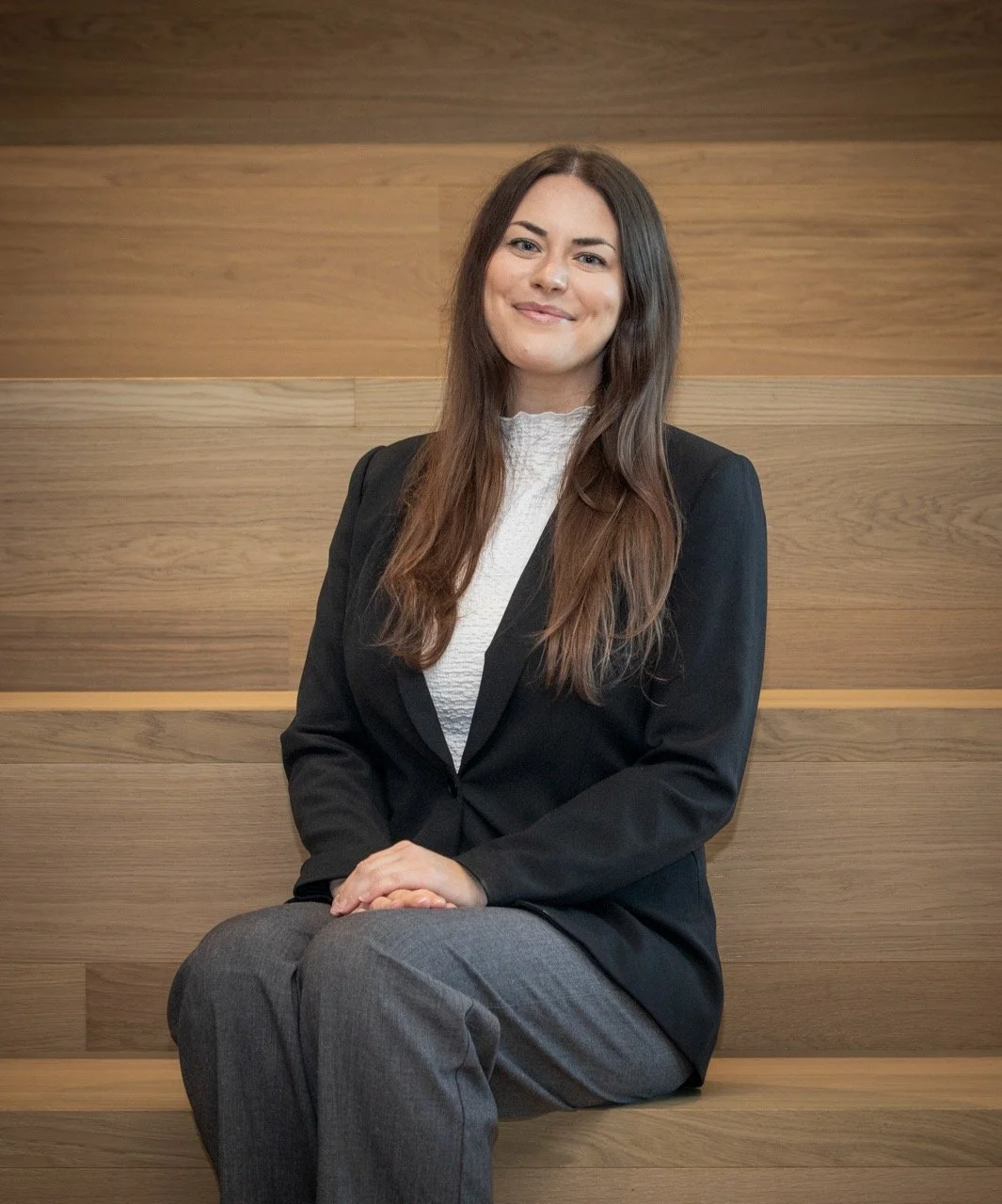 Dame med lang brunt hår, iført svart blazer og grå bukse, sitter på en trebenk med en trevegg i bakgrunnen.