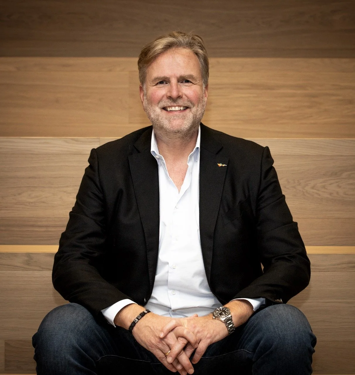 Mann i svart blazer og hvit skjorte sitter på en stol med en trevegg i bakgrunnen.
