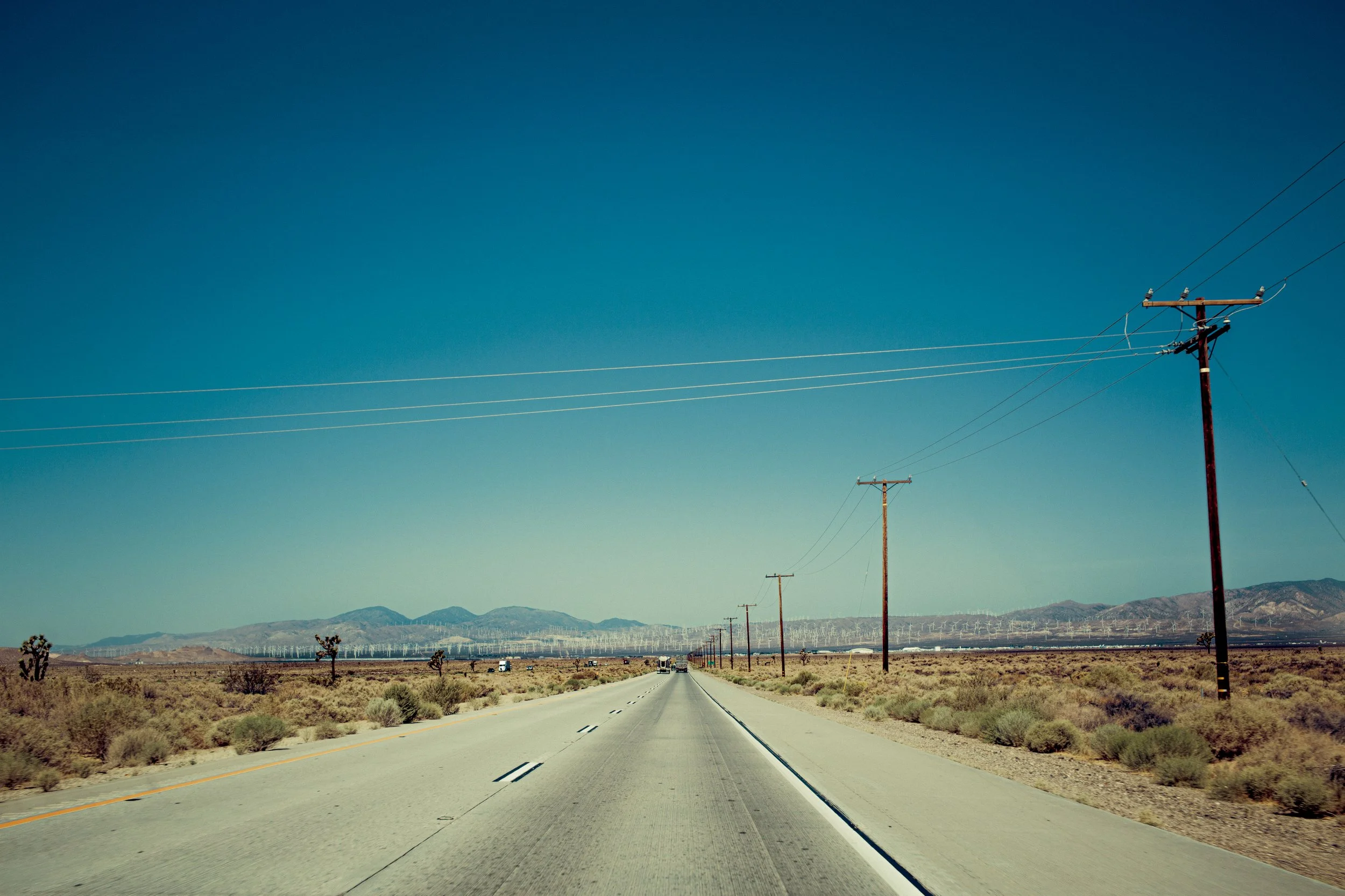Une route droite traversant un paysage désertique avec des montagnes en arrière-plan, des lignes électriques sur la droite et le long de la route, sous un ciel dégagé et bleu.
