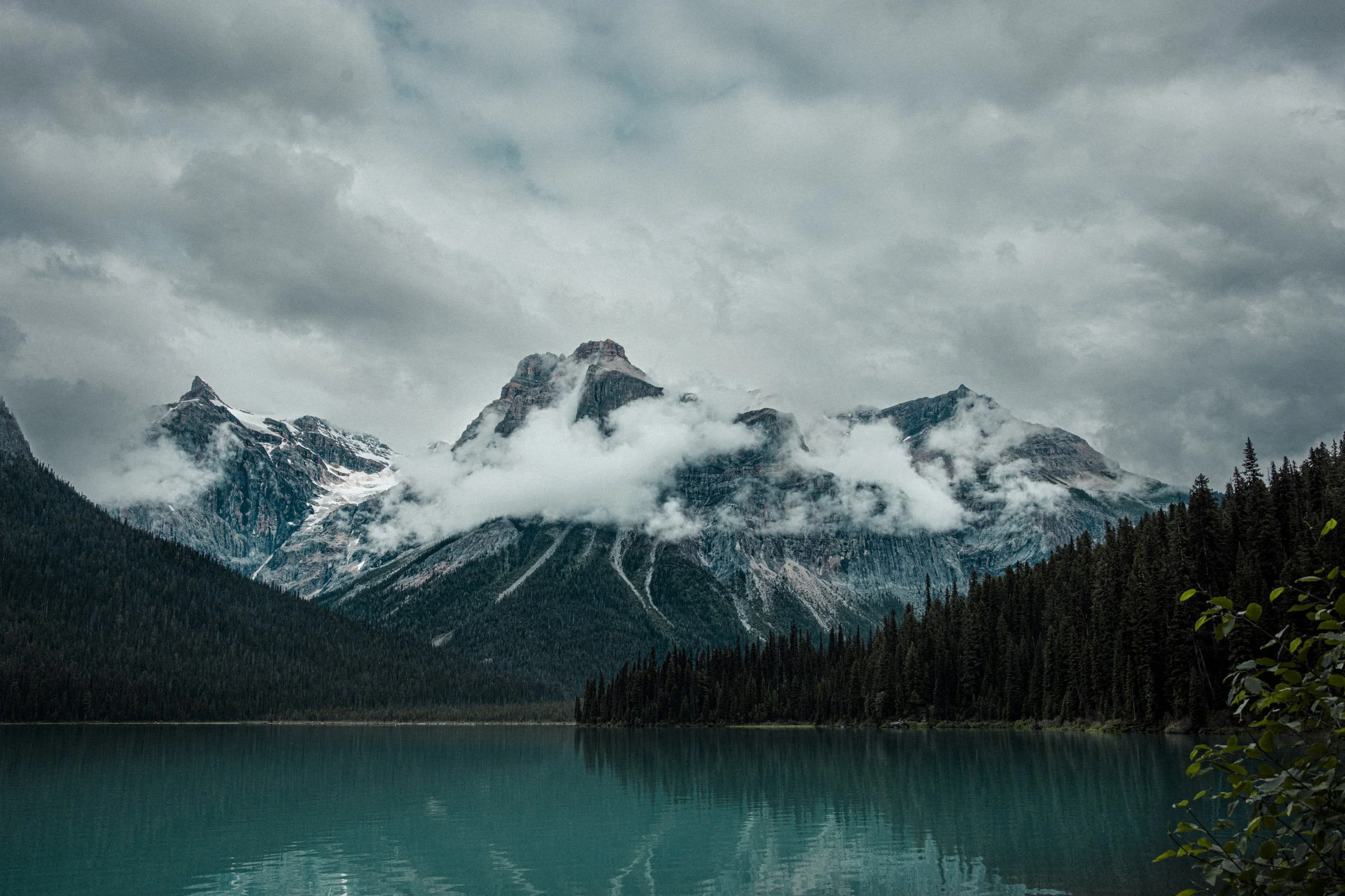 Montagnes enneigées et brumeuses au bord d'un lac turquoise, ciel nuageux.
