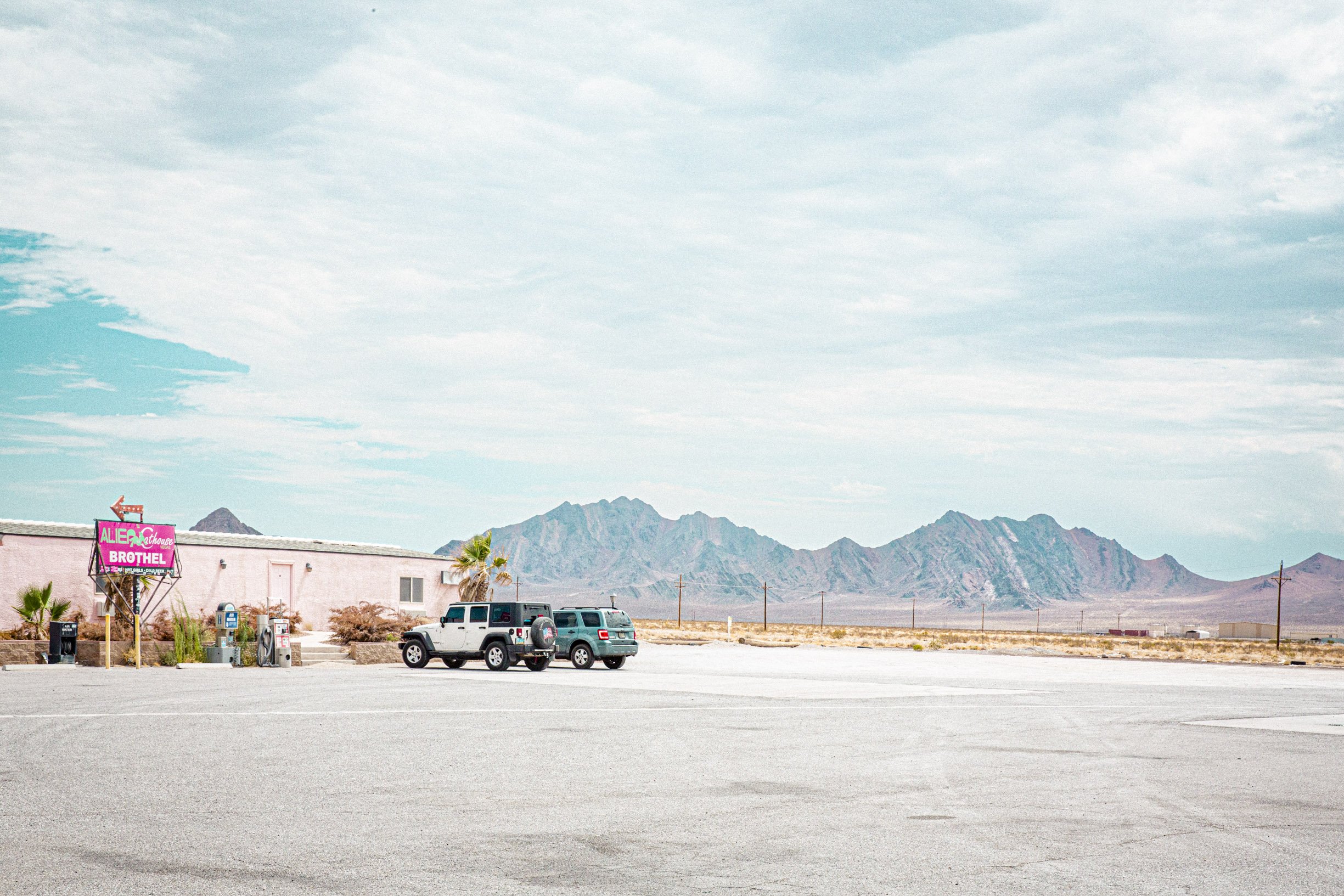 Un parking désert avec quelques voitures, une station-service, un bâtiment avec un panneau rose et vert, des palmiers, un paysage désertique et des montagnes au loin, sous un ciel nuageux.