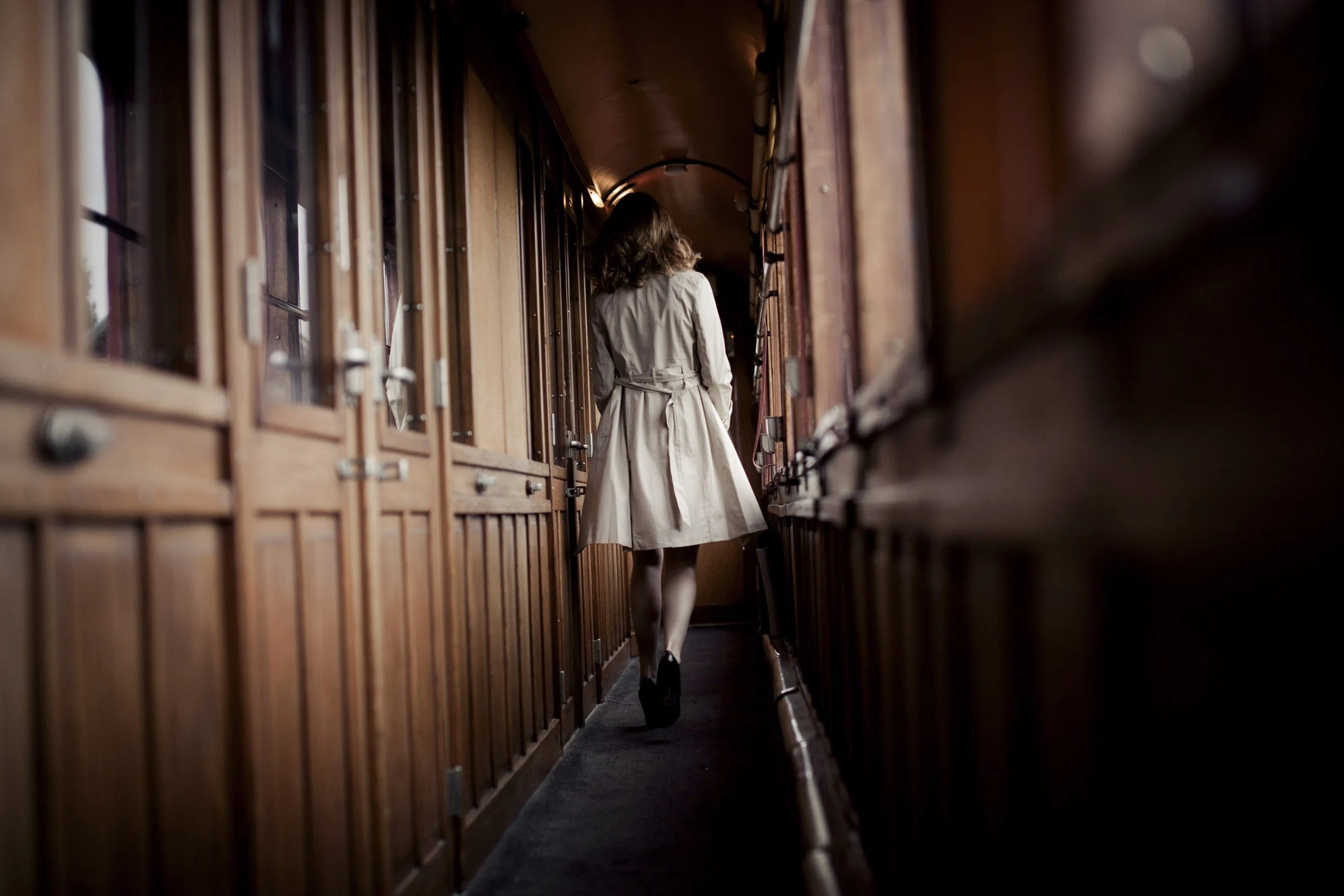 Une femme portant un trench beige marche dans un couloir étroit et sombre entouré de boîtes en bois ou en métal.